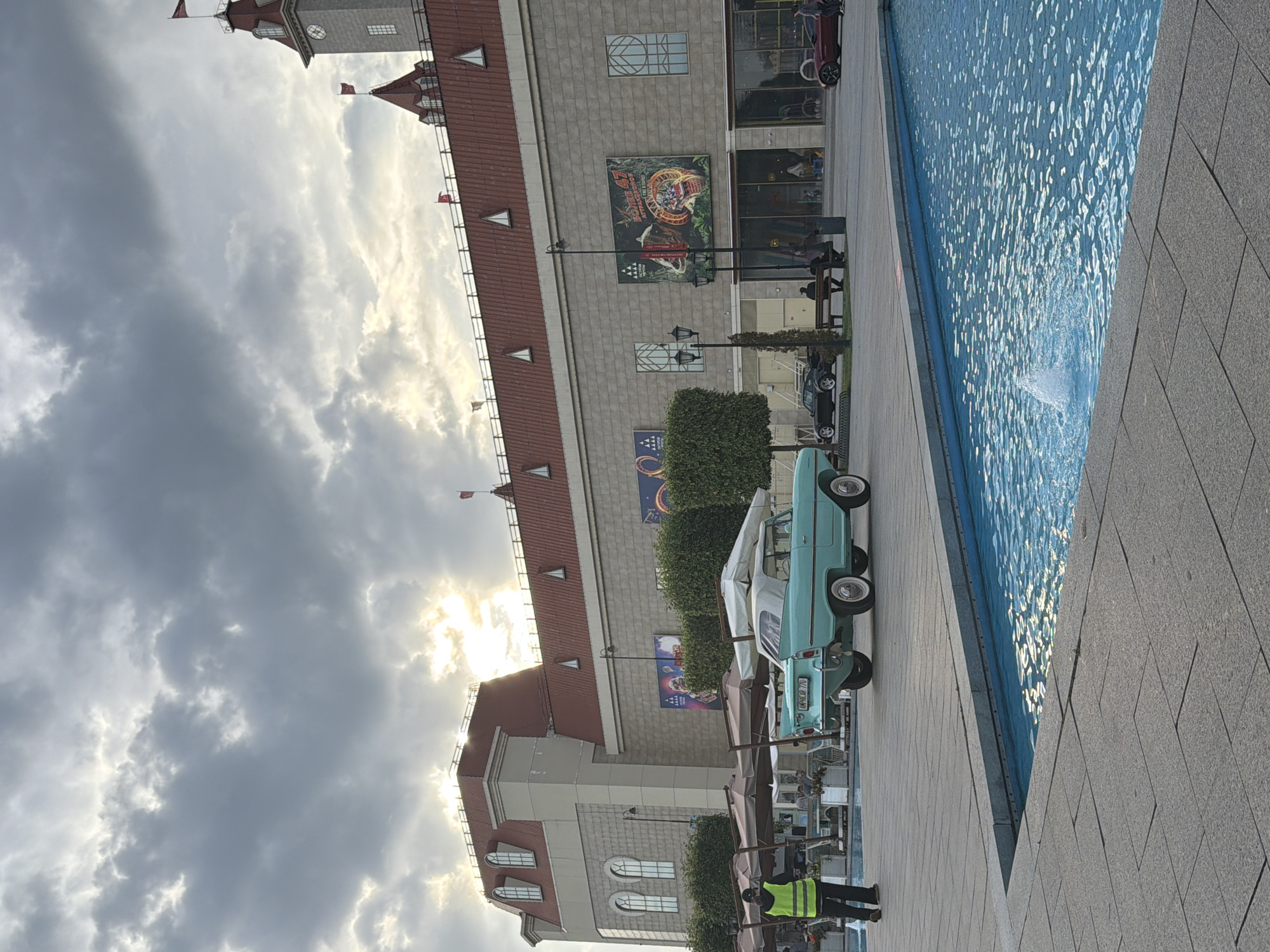 In an outdoor plaza, a light blue amphibious car is parked near a fountain. The car has a white convertible top and is facing away from the viewer, with its rear end visible. The fountain's water is clear blue and has a small jet of water spraying upwards. In the background, a large, light-colored building with a red-tiled roof and a clock tower is visible. The sky is cloudy, with patches of sunlight breaking through. A person wearing a high-visibility vest is standing near the left side of the image, facing away from the car. Several umbrellas are set up in front of the building, and some people are seated under them. Other cars are parked in the background, including a black sports car and a red sports car. The plaza is paved with large, gray stone tiles. The image appears to be taken in the daytime.