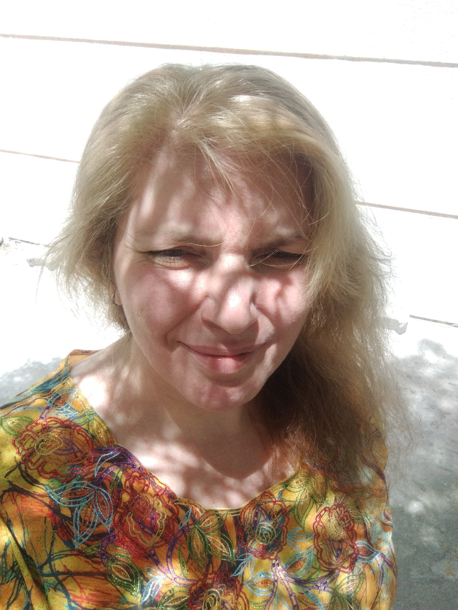 A medium shot captures a fair-skinned woman with blonde hair and a slight smile. Her hair is somewhat disheveled, and shadows from foliage fall across her face. She is wearing a colorful, floral-patterned top. The background appears to be a white wall. The location is in the city of Al Mas'oudi, United Arab Emirates.
