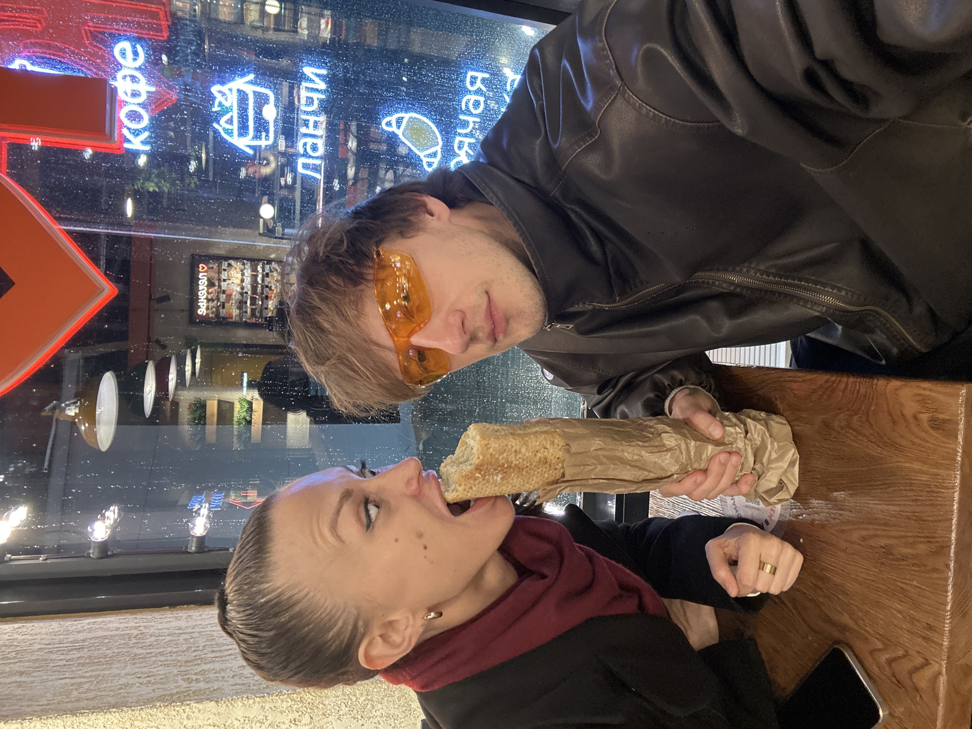 A photograph shows a woman biting into a baguette while a man holds the baguette in front of her mouth. The man is wearing a brown leather jacket and yellow-tinted sunglasses. The woman is wearing a black coat and a burgundy scarf. They are sitting at a wooden table, on which there is a smartphone. A large window is behind them, with neon signs visible. The neon signs have text in Russian. One sign reads "кофе" (coffee). The reflection in the window also includes street lamps and other buildings. The neon signs, including the text "ДАНАЧИ" and an image of a croissant, suggest the photograph was taken in Murino, Russia. It is raining outside.