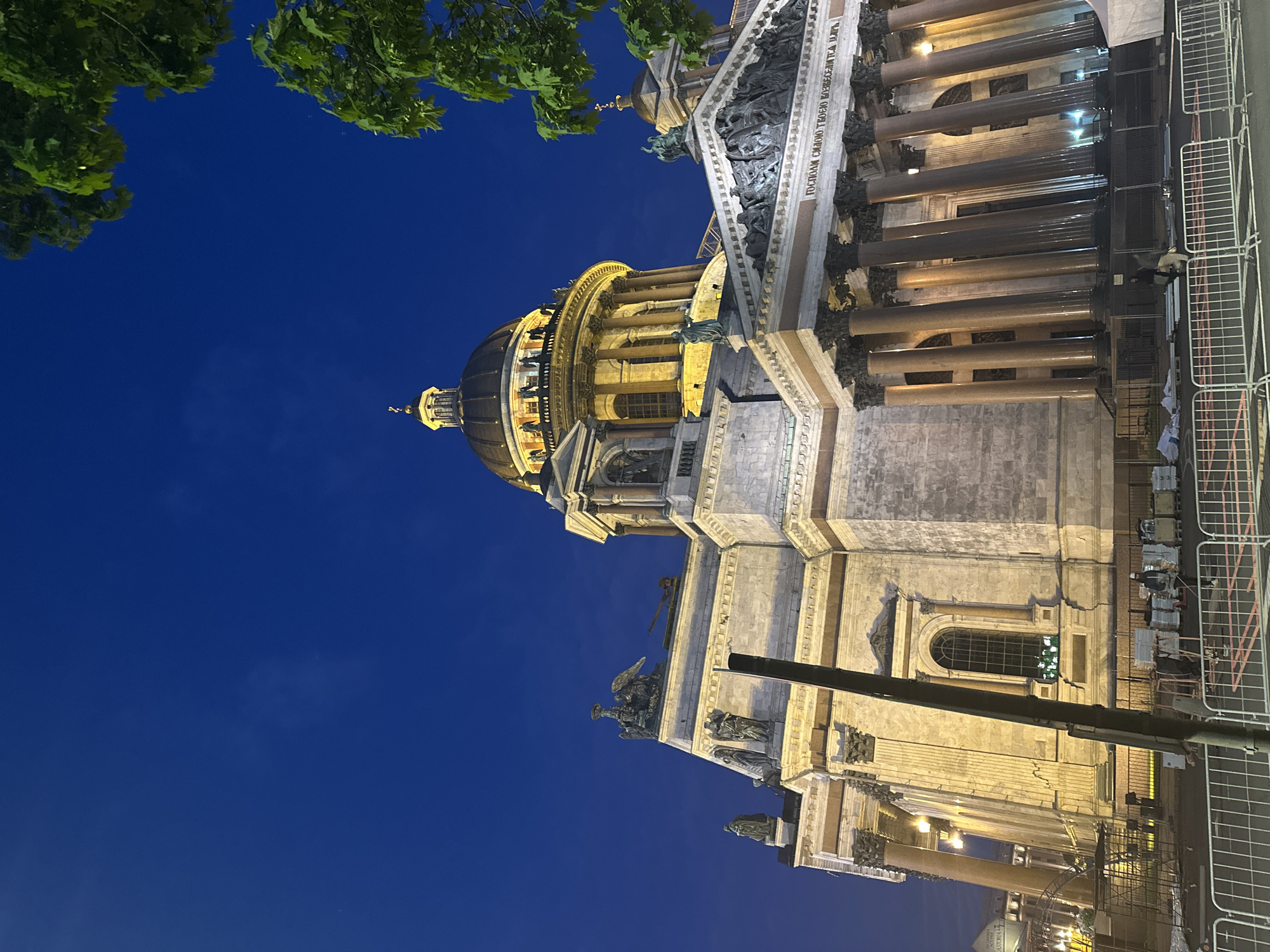 Here's a description of the image:
The image showcases a section of a large, ornate cathedral against a deep blue evening sky. The architecture is characterized by classical elements, including prominent columns, sculptures, and intricate details along the facade. The building appears to be illuminated by artificial lights, highlighting its architectural features. A large dome with a golden top is visible on the left side of the structure. In the foreground on the right side, there is a section of construction fencing, suggesting ongoing maintenance or restoration work on or around the building. The top left corner of the image includes some tree foliage. The image appears to be taken from a lower angle, looking up at the building.