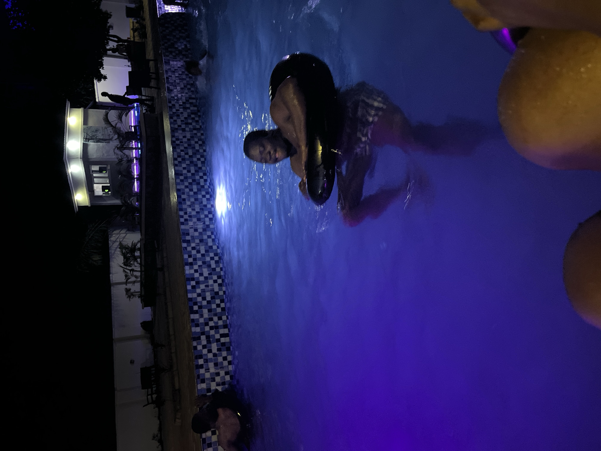 In Kason, Nigeria, a man is pictured at night in a swimming pool, using a black inflatable tube as a float. The pool is illuminated by blue and purple underwater lights, casting a vibrant glow on the water. The man, wearing patterned swim trunks, is looking towards the camera with a smile and his arms are resting on the tube. In the background, a building with bright lights is visible, along with some vegetation and a white wall surrounding the property. Other individuals are present in the pool, some partially submerged. The scene suggests a leisurely evening at a pool, likely a resort or a private residence.