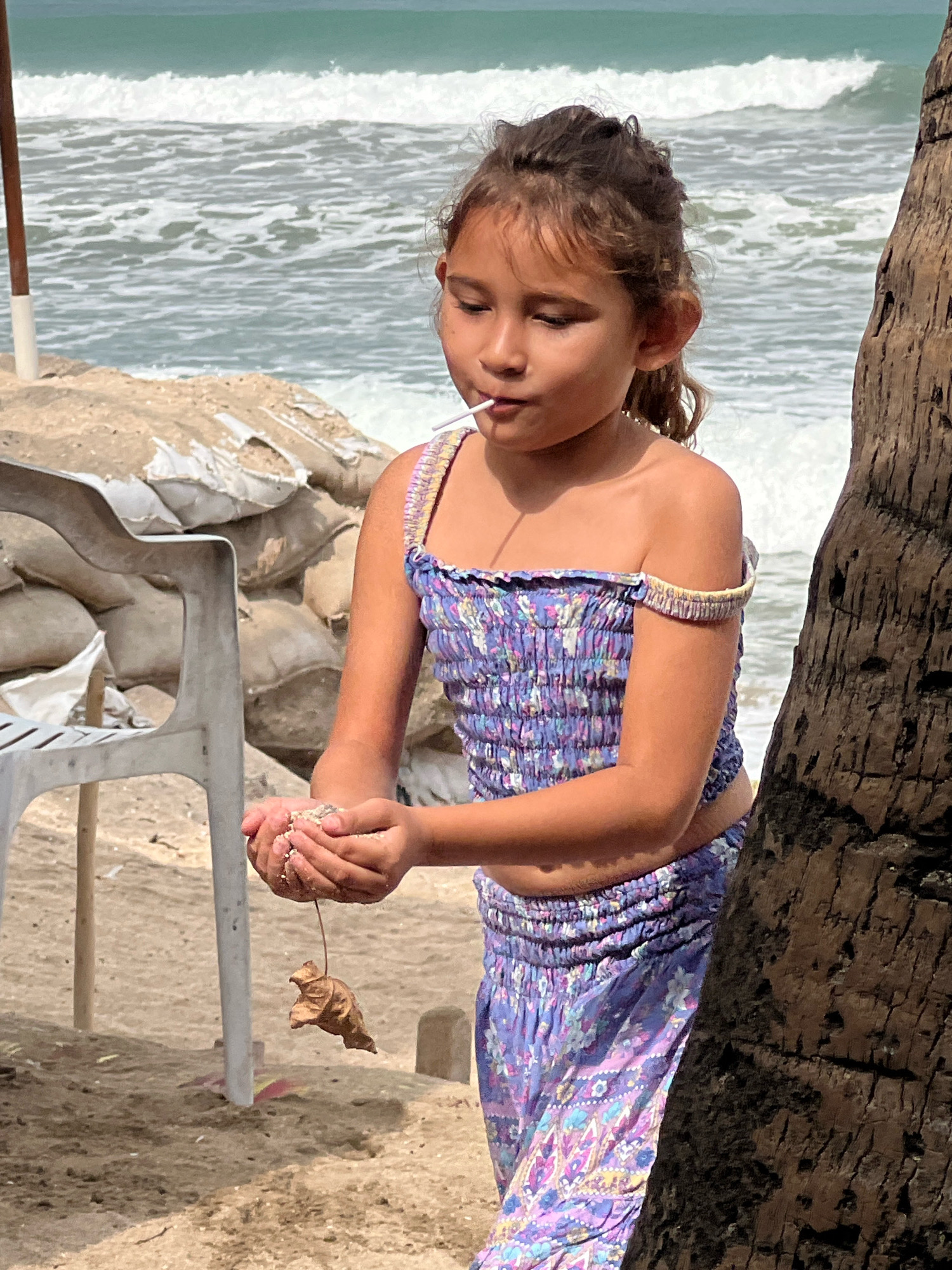 A young girl stands on a sandy beach, partially obscured by a palm tree trunk on the right. She is wearing a purple floral-patterned two-piece outfit, with her hair pulled back in a bun. The girl holds a small amount of sand in her hands, and a dried leaf hangs from a string attached to her fingers. She has a lollipop in her mouth. In the background, the ocean stretches out with waves breaking on the shore under a bright, sunny sky, suggesting it is daytime with clear weather. Behind her, there are sandbags and a white plastic chair, hinting at a possibly temporary or protected area on the beach. The overall scene captures a moment of quiet observation or play by the sea.