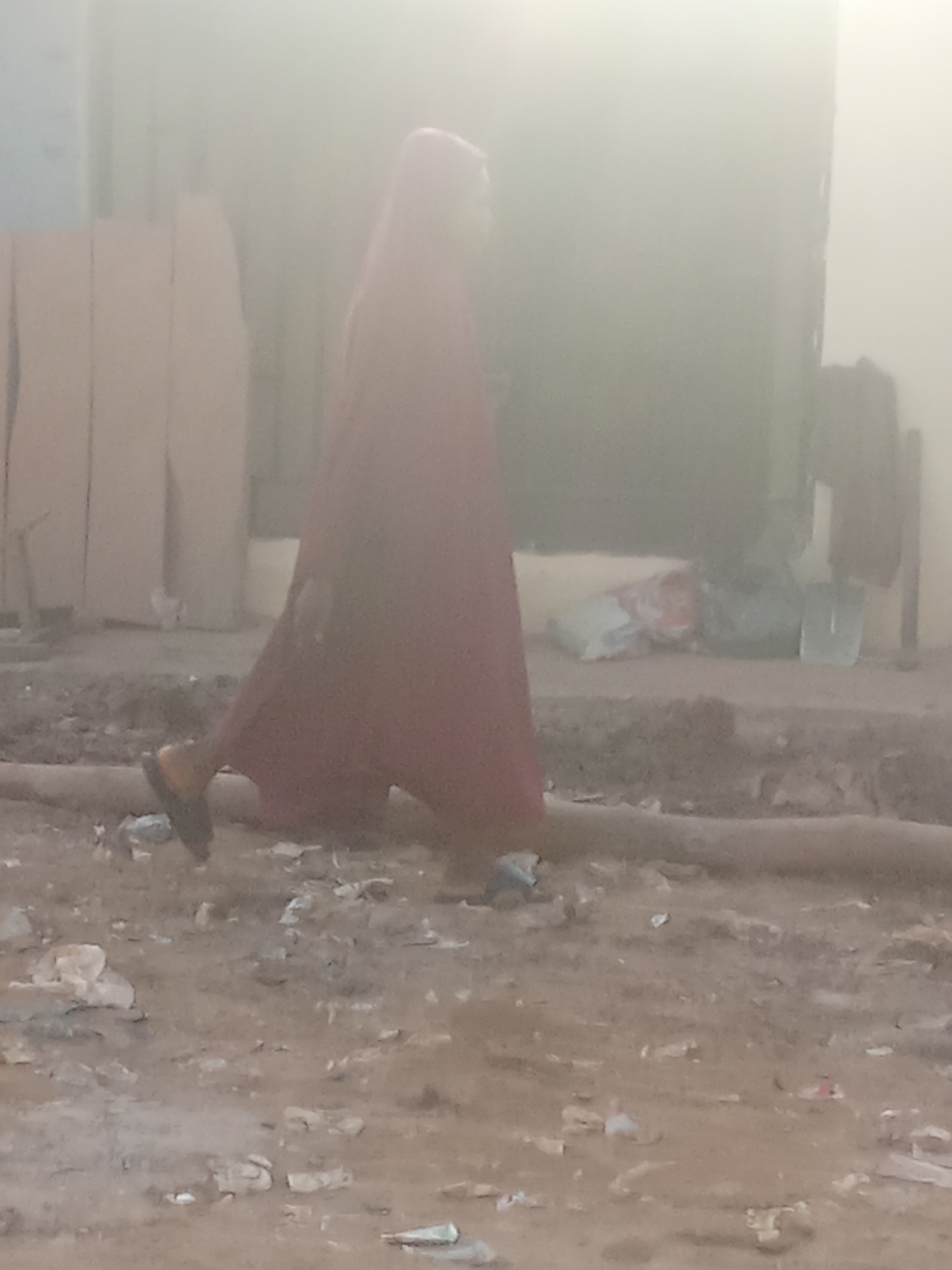 The image depicts a person walking in Kagini, Nigeria, on a dusty ground with scattered debris. The individual, possibly a woman due to the red garment covering their head and body, is walking from left to right, with their left foot appearing to be shod in a dark sandal. The background is out of focus and hazy, but suggests a built environment with what appear to be large, light-colored rectangular structures on the left, possibly stacks of materials or construction elements. To the right of the person, in the middle ground, are several objects including bags or sacks and a shovel leaning against a vertical structure. The ground is uneven and appears to be a mixture of dirt, gravel, and small stones, with various pieces of trash, such as plastic bags and paper, scattered throughout. A cylindrical object, possibly a pipe or hose, runs horizontally across the lower part of the image. The overall lighting is diffuse, suggesting an overcast day or dusty atmospheric conditions.
