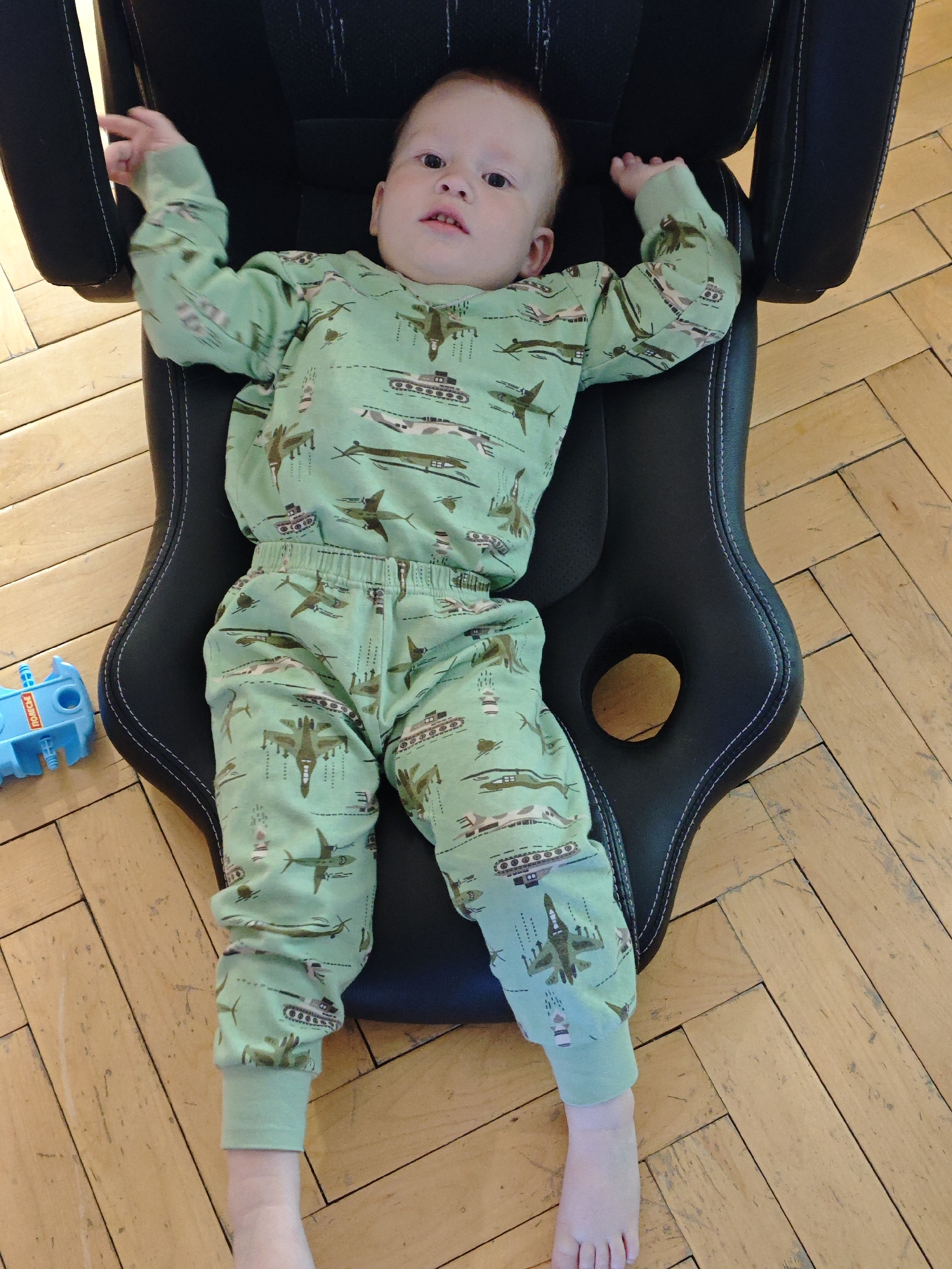 A baby wearing green pajamas patterned with tanks and airplanes is lying in a black gaming chair on a herringbone-patterned wooden floor. A blue toy is on the floor near the chair. The location cannot be determined from the image.