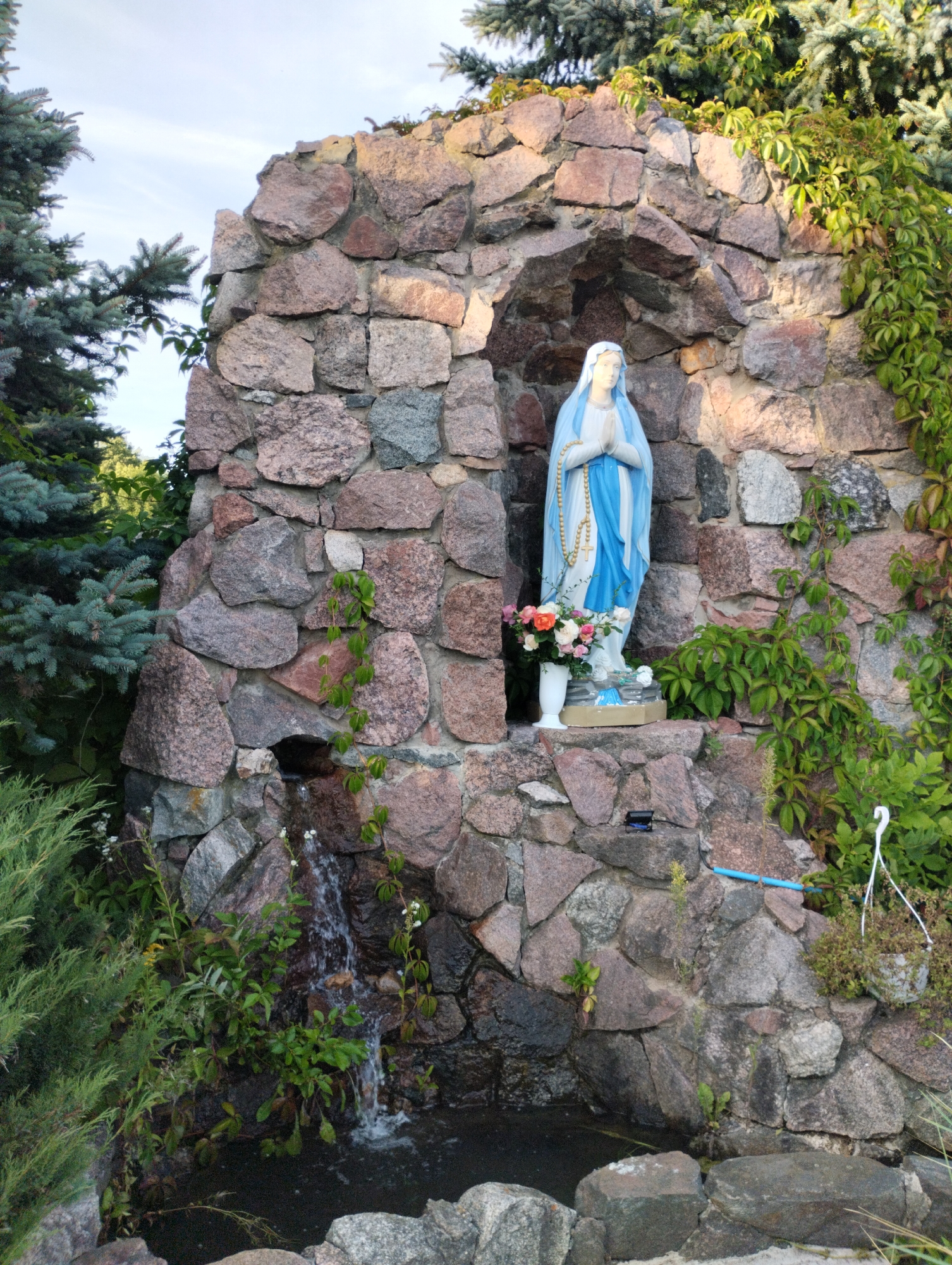 In a grotto constructed of rough-hewn stones, a statue of the Virgin Mary, draped in a blue and white robe and holding a rosary, stands with her hands clasped in prayer. A small waterfall cascades down the stone structure into a pool below. The grotto is adorned with greenery, including a climbing vine and surrounding evergreen trees, suggesting an outdoor setting. The statue is positioned on a rocky ledge, with a vase of flowers placed beside it. A small, dark object, possibly an electronic device, is visible on the rock ledge to the right of the statue. The scene is set within the natural landscape of Вішнеўскі сельскі Савет, Belarus.
