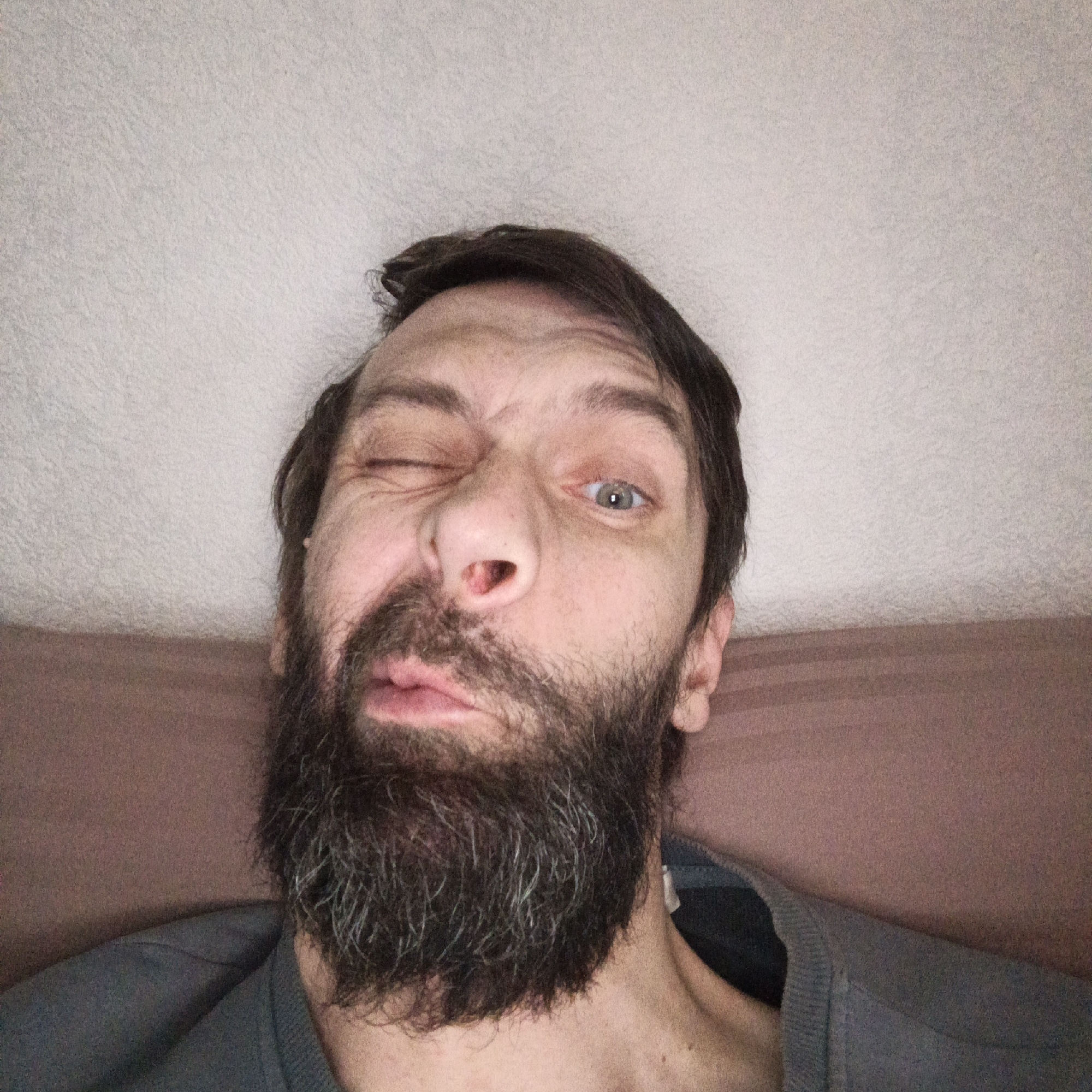 A man with a beard and dark hair is making a silly face. He is winking his left eye and distorting his mouth. The background is a white ceiling and a brown wall. The location of the scene is unknown.