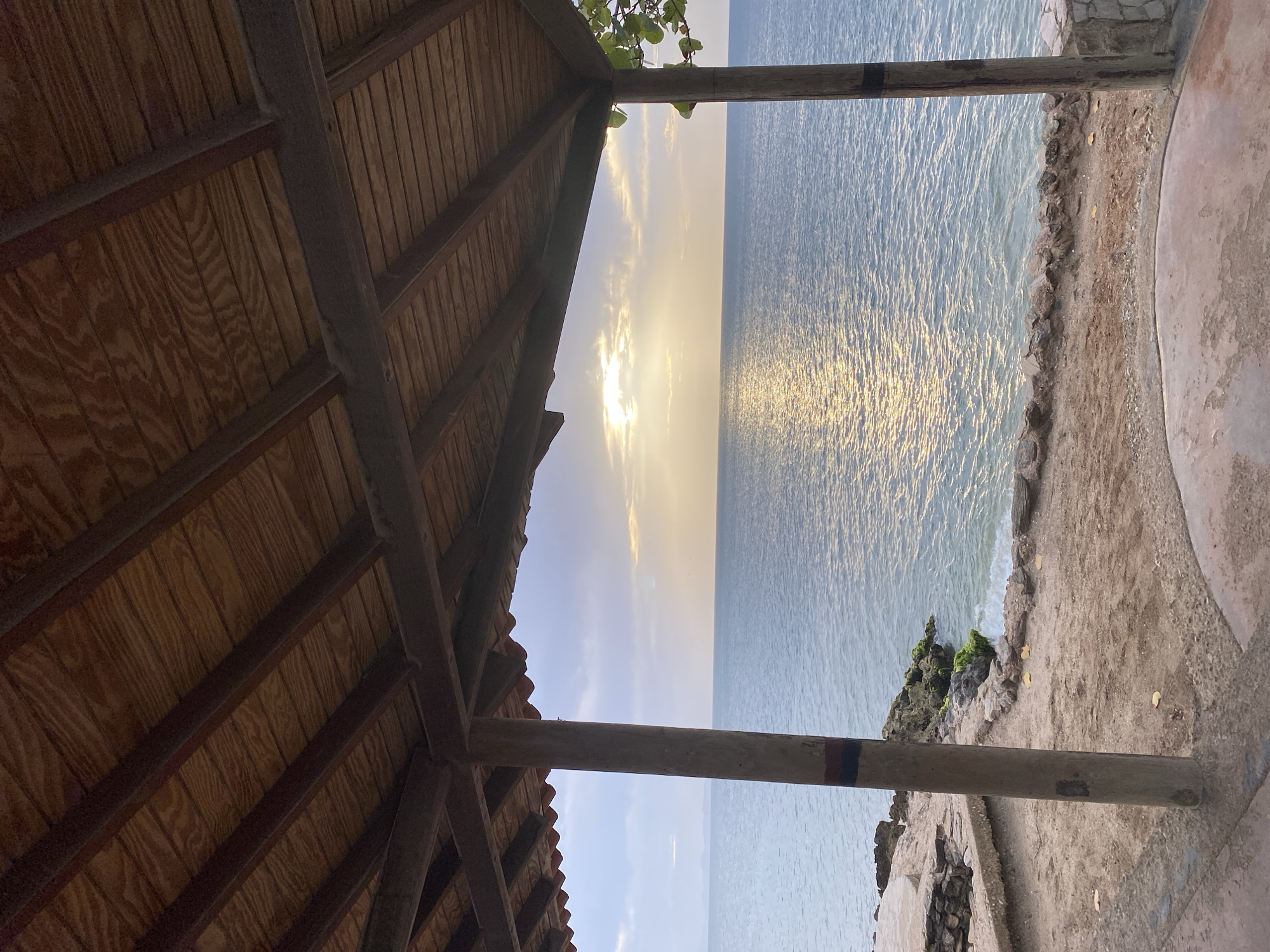 The image shows a view of the sea from under a wooden-roofed structure. The structure has visible wooden beams and planks forming the ceiling. In the foreground, there are two wooden posts, one on the left and one on the right, supporting the roof. The post on the left has a red and blue stripe painted around its lower section. The ground beneath the structure is a mixture of sand and concrete.
Beyond the structure, a calm sea stretches to the horizon. The sun is low in the sky, casting a golden reflection on the water. The sky is partly cloudy, with the sun peeking through the clouds. On the left side of the image, there are rocky outcrops along the shoreline, some of which are covered in green algae. On the right side, there is a glimpse of green foliage.
This scene is likely located in Juan Griego, Venezuela, a coastal town known for its beaches and scenic views of the Caribbean Sea. The presence of the simple, open-air structure suggests it might be part of a beachside establishment, a public viewpoint, or a private residence with a view of the ocean. The overall atmosphere is peaceful and serene, indicative of a sunset or sunrise by the sea.
