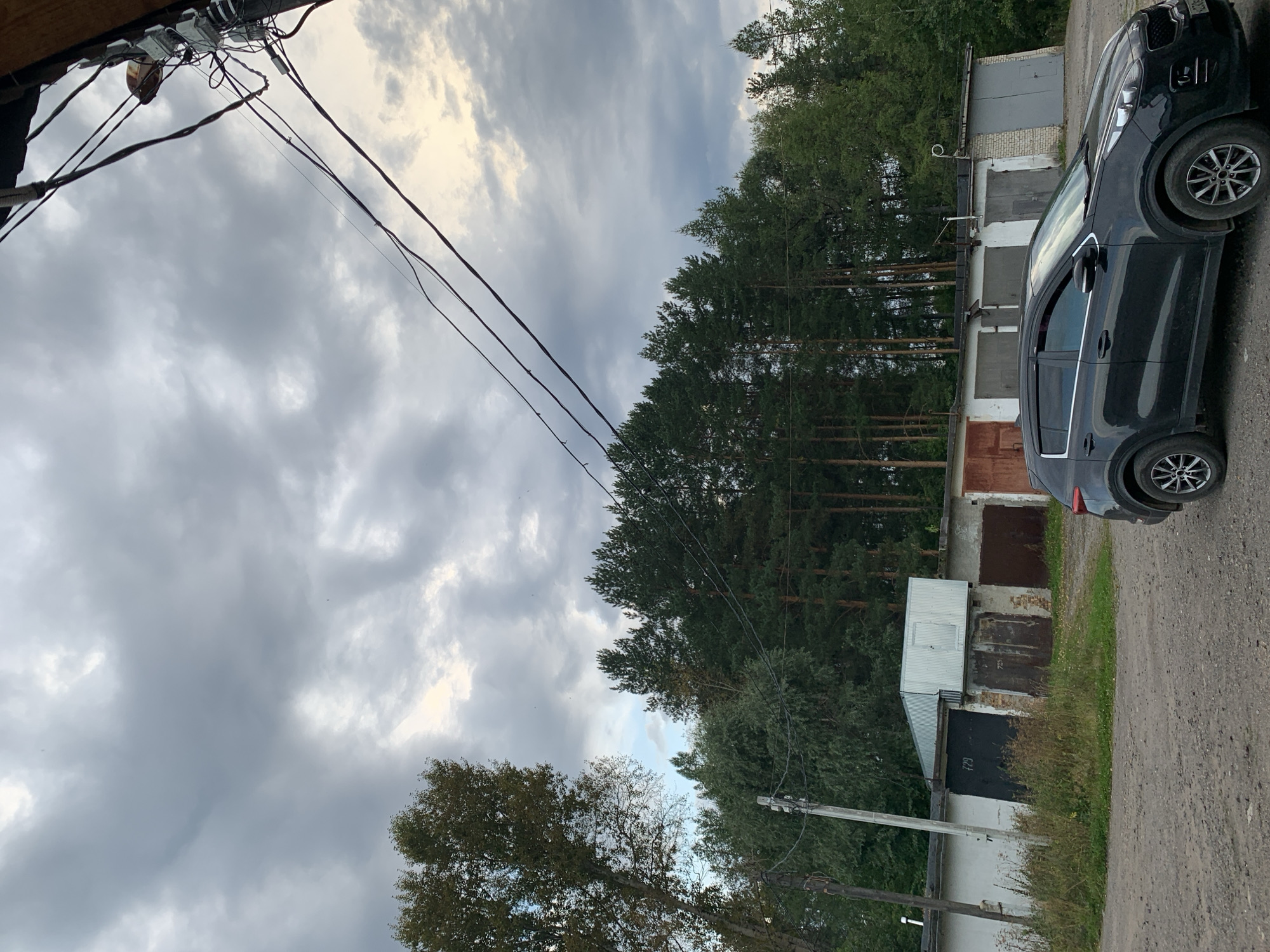 A dark grey SUV is parked on a gravel road in front of a row of garages with corrugated metal doors. Some of the garages have numbers on them, such as "729". The garages are lined up against a white wall with some brickwork visible. Behind the garages, there is a dense forest of pine trees. The sky is cloudy and overcast, with some hints of light breaking through. A utility pole with wires runs across the frame in the foreground and over the garages. The scene appears to be an urban or suburban setting, likely in a garage cooperative or industrial area, given the presence of multiple garages and utility infrastructure. The overall impression is one of a slightly overcast, late afternoon or early evening in a sparsely populated area.
