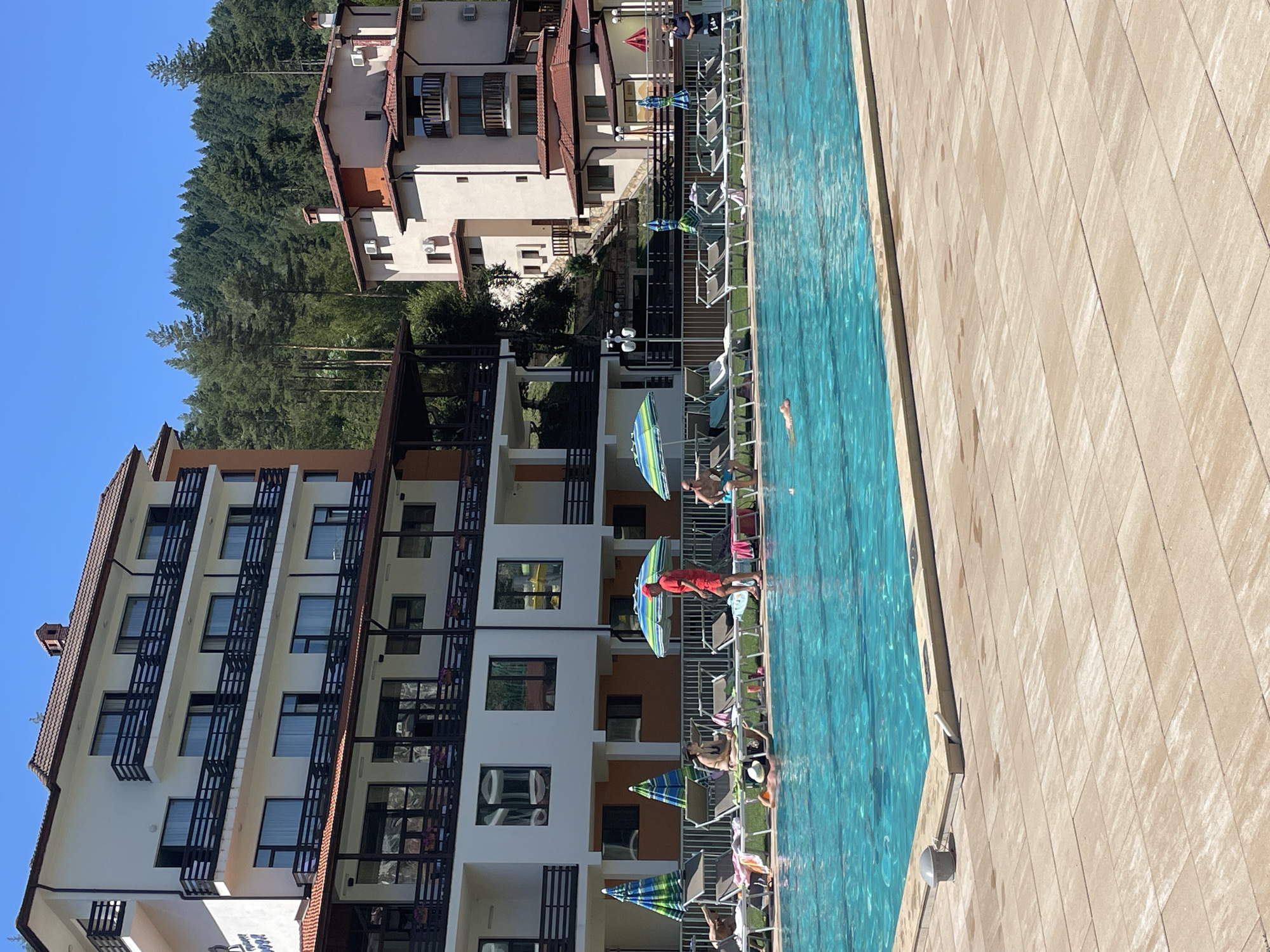 In Banite, Bulgaria, a scene unfolds by a bright blue swimming pool. On the left, a large, white hotel with multiple balconies and dark railings stands against a backdrop of a dense, green pine forest under a clear blue sky. To the right of the hotel, another similarly styled building with light-colored walls and terracotta roofs is visible, also nestled against the treeline.
The pool area is furnished with numerous lounge chairs, many occupied by people. Some are sunbathing on the chairs, while others are relaxing poolside. Three individuals are in the water; one is actively swimming, while two others appear to be submerged in shallow areas. Umbrellas are dotted around the pool deck, providing shade.
On the far right of the scene, a man in a blue shirt stands by the edge of the pool, observing the activity. Near the pool, a person in a red shirt and red shorts stands, appearing to be a lifeguard or a staff member. The pool deck is paved with light-colored rectangular tiles. The overall atmosphere is one of leisure and relaxation on a sunny day.