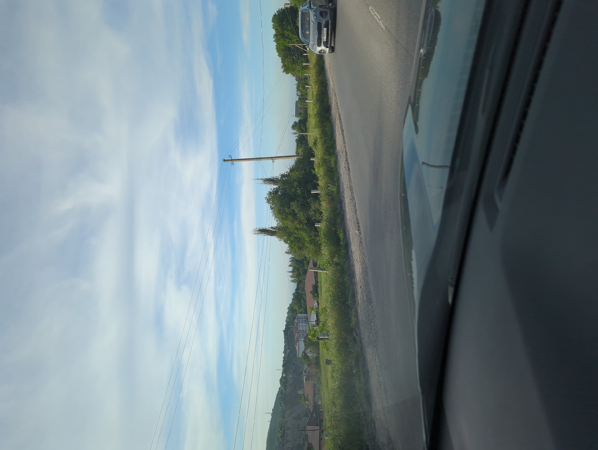 Captured from inside a car, the photograph shows a road winding through Beş-Küŋgöy, Kyrgyzstan. Another car is visible ahead on the road. Greenery borders the road, extending into the background where houses and buildings can be seen. Utility poles and wires run alongside the road. The sky is visible above, showing a mix of blue and clouds.