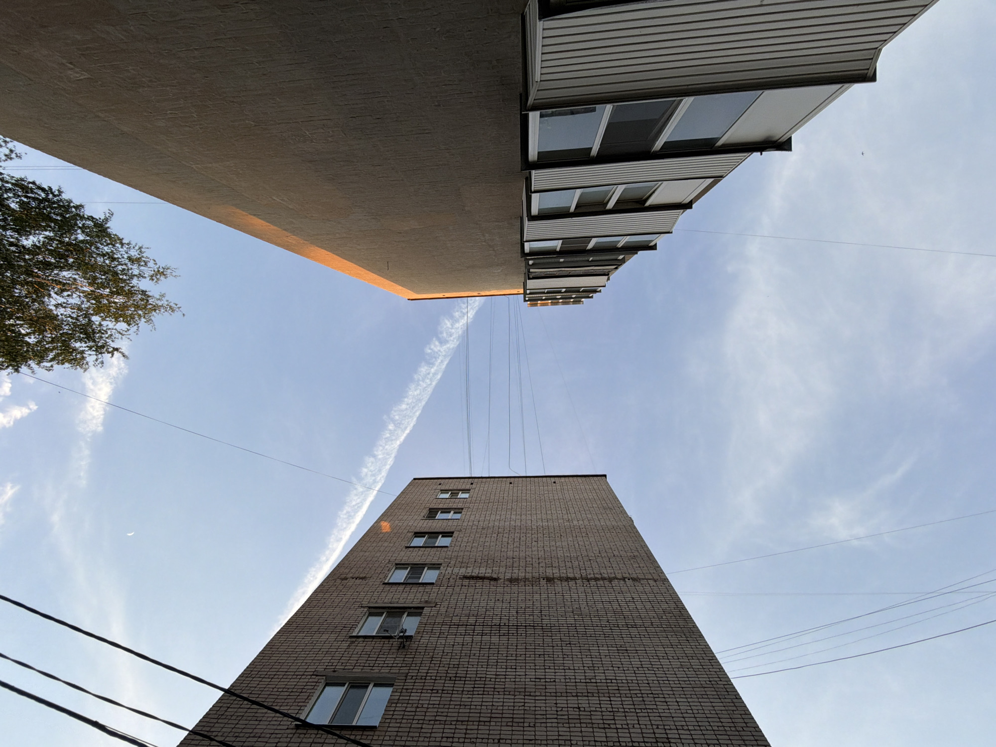 The image is a low-angle shot looking upwards in what is reported to be the city of Chaikovsky, Russia. It captures two buildings and a portion of the sky. One building, constructed of brick, rises toward the bottom of the frame and features multiple windows. The other building, with visible white panels and windows, appears towards the top. Electrical wires stretch across the open sky, and a faint contrail is visible. There is also a partially visible tree on the left side of the image. The sky is pale blue with light cloud cover. The lighting suggests either early morning or late afternoon. No people or animals are visible.