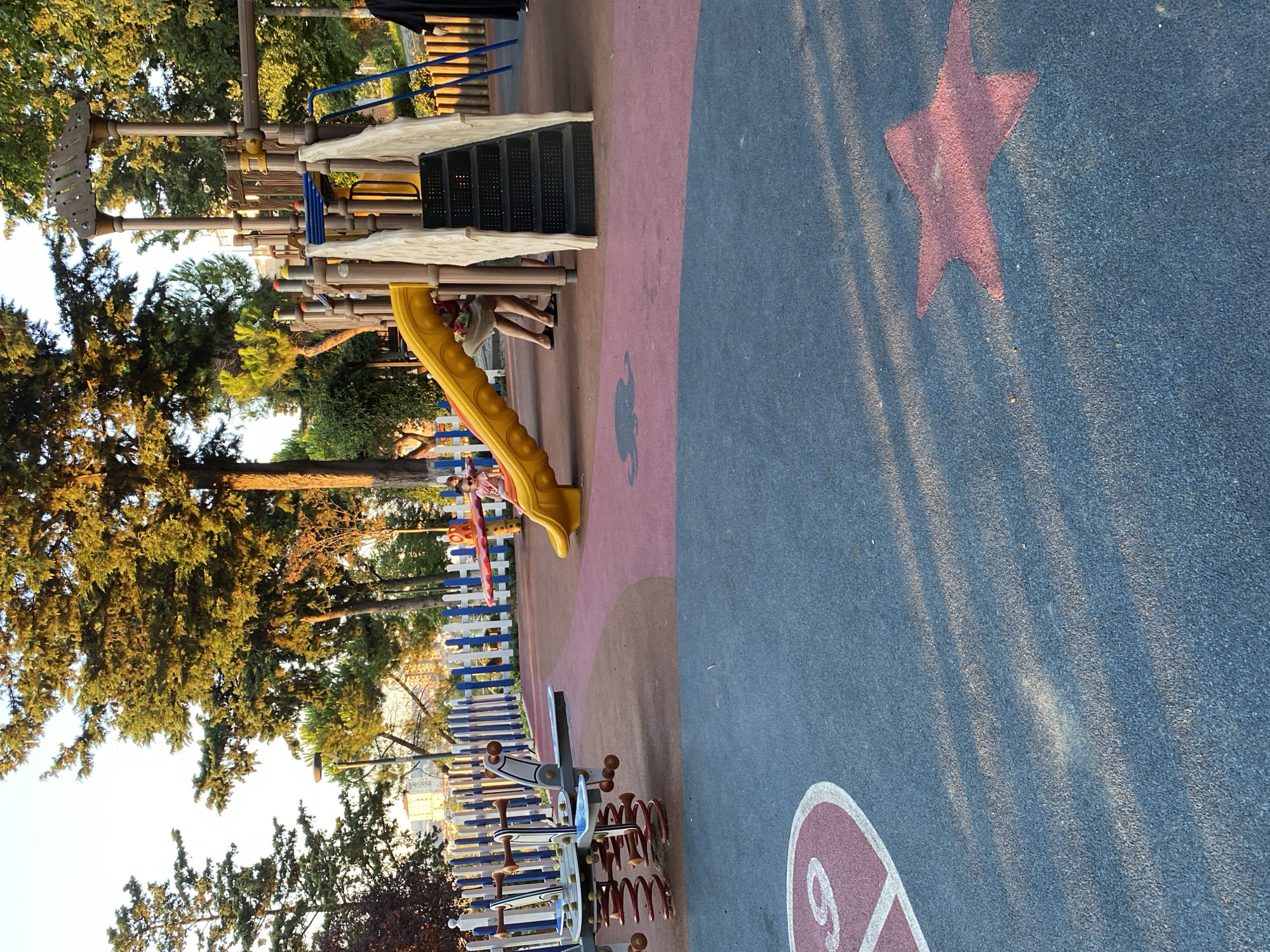 An outdoor playground in Küçükçekmece, Turkey, is depicted in the image under a clear sky, with dappled sunlight filtering through surrounding trees. The playground features a colorful climbing structure with a yellow slide and stairs. A child is visible on the slide, wearing a pink shirt and sunglasses. Nearby, adults are present, one of whom is partially obscured by the structure. In the foreground, the playground surface is a mosaic of red and blue material, marked with white designs of a heart and a star. A spring rider toy is also situated in the left portion of the scene, with a white and blue fence bordering the playground in the background.