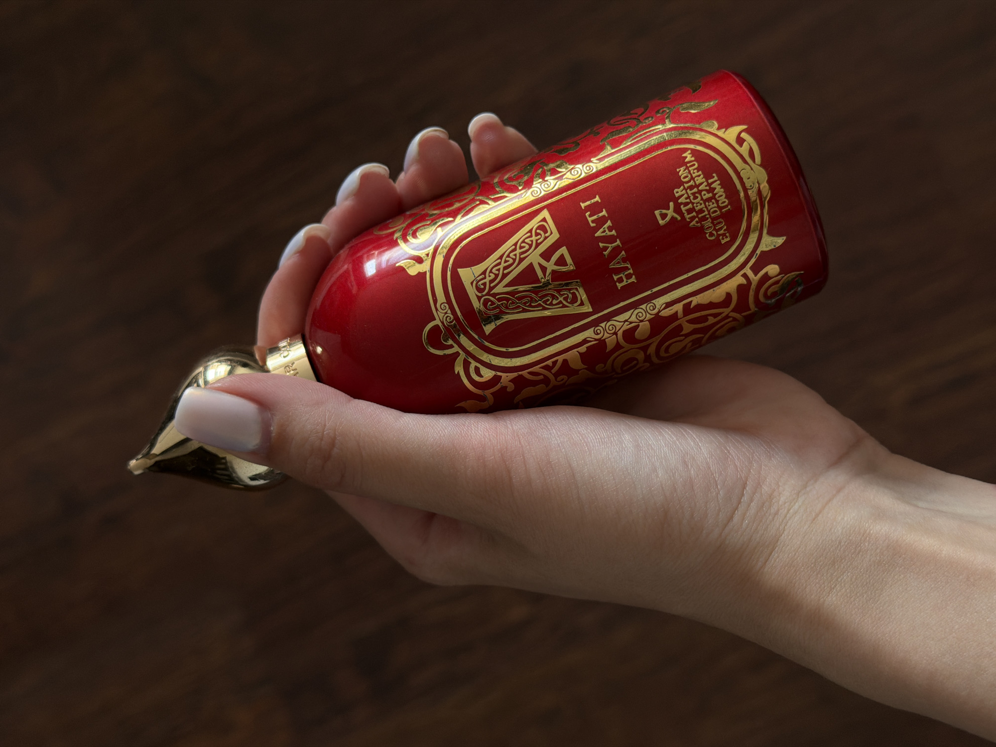 A hand is holding a red perfume bottle with gold accents. The bottle has the word "HAYATI" written in gold lettering, as well as other gold decorative elements. The background is a dark, wooden surface. The scene is a close-up shot, with the focus on the perfume bottle and the hand holding it.