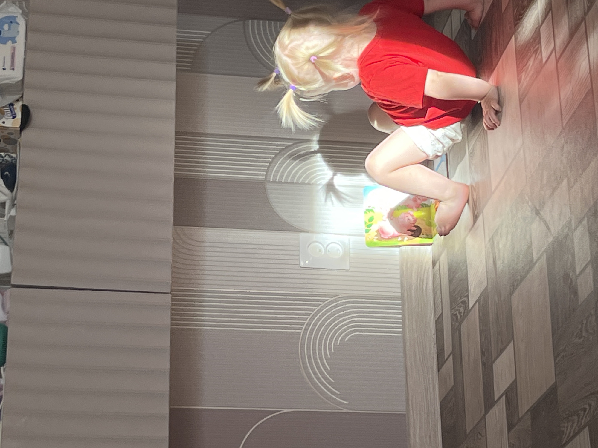 A toddler with blonde hair in pigtails, wearing a red shirt and white shorts, is on the floor in front of a wall with a geometric pattern. The toddler is facing away from the camera, looking towards a bright light source that is illuminating an object on the floor. The object appears to be a toy or a package with colorful imagery. The floor is tiled with a wood-grain pattern. The scene takes place indoors, likely in a residential setting. The light casts shadows on the wall, creating an interesting visual effect. The location is identified as Rakhinka, Russia, but there are no visual cues in the image to confirm this specific location.