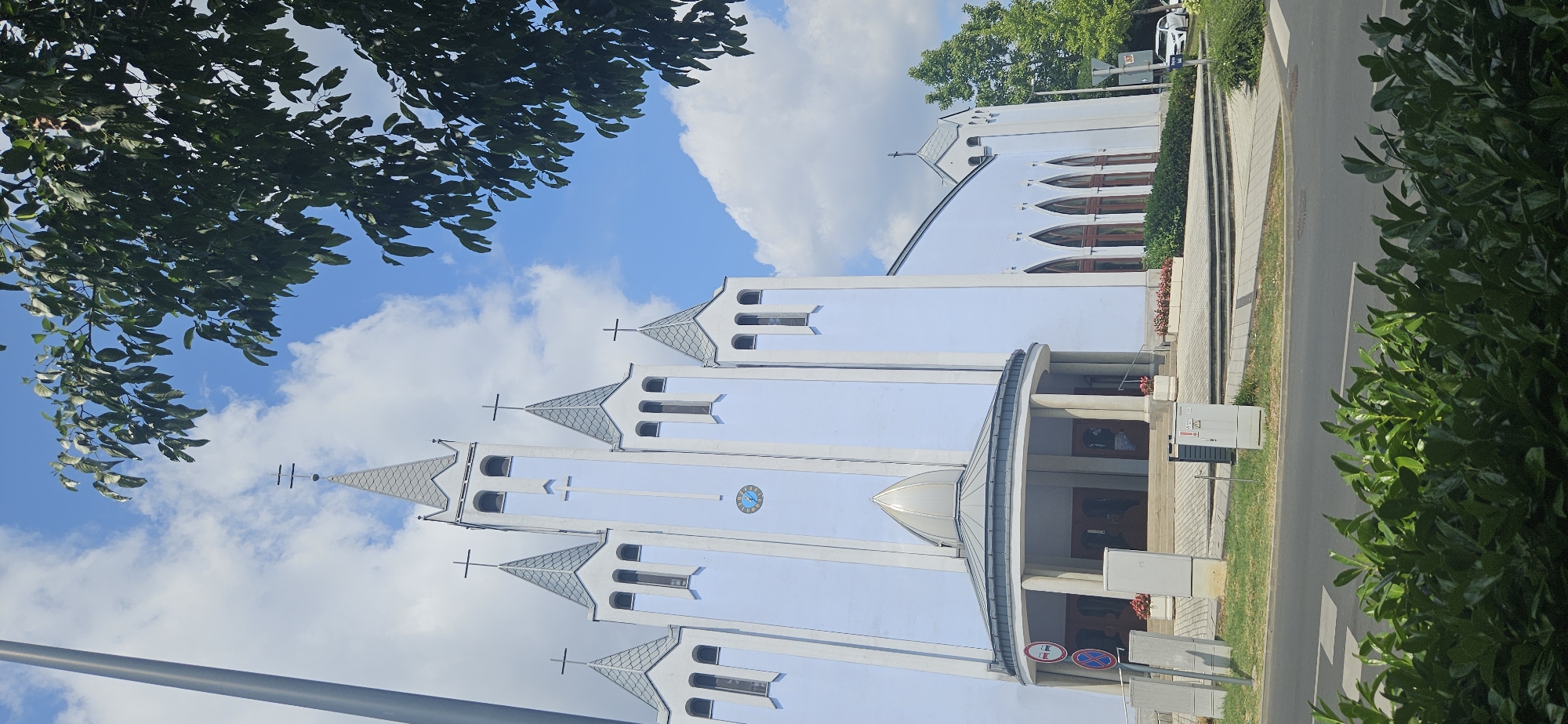 This is a vertical, eye-level, medium shot of a modern, light blue church building in Hévíz, Hungary. The church has a central facade with five prominent towers, each topped with a pointed, grey roof and a cross. The central tower is the tallest and features a clock face. The entrance to the church is a circular, covered porch supported by columns, with a domed roof. The building's walls are smooth and light blue, with grey accents on the towers and roofs. To the right of the church, there are several tall, arched windows with dark brown frames. The building is situated on a paved area with steps leading up to the entrance. In the foreground, there is a road with a crosswalk, and lush green bushes line the edge of the road. A tall, grey utility pole stands to the left of the church. The sky is blue with scattered white clouds, and the upper branches of a large tree with dark green leaves are visible in the top right corner of the frame. The scene appears to be calm and undisturbed, with no people or overt activity visible.