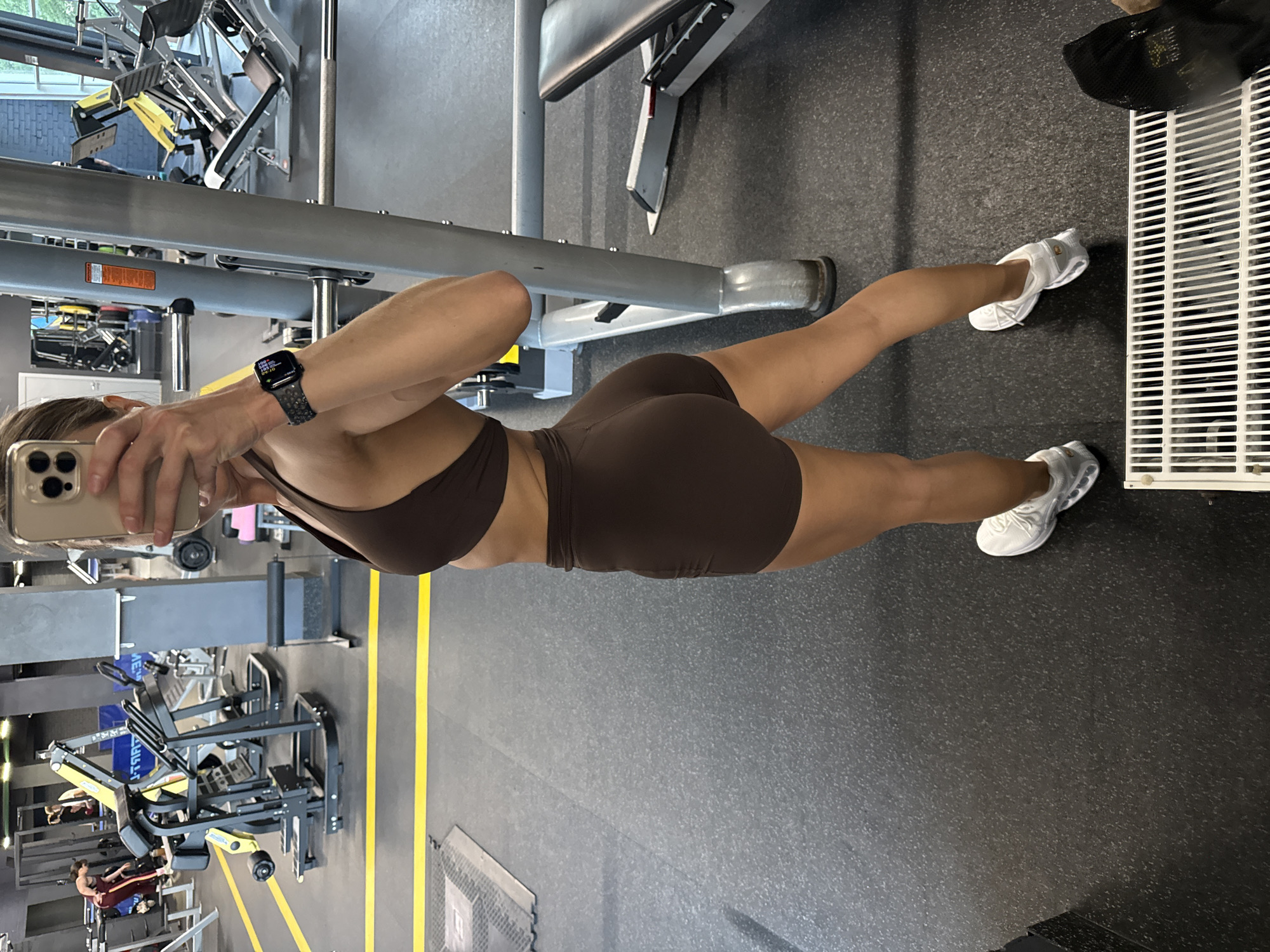 A female individual is captured in a mirror selfie within a fitness gymnasium in Saint Petersburg, Russia. The subject is positioned facing slightly away from the camera, looking over her left shoulder into the smartphone she holds up with her right hand. She is wearing a dark brown two-piece athletic ensemble consisting of a sports bra and form-fitting shorts. An Apple Watch with a dark, textured band is present on her left wrist, displaying digital information. Her hair is pulled back, and white athletic shoes with gray accents are visible on her feet.
The gym environment features dark gray rubberized flooring with yellow demarcation lines. In the background, various pieces of strength training equipment are visible, including multiple leg press machines (yellow and black), a large black and yellow machine (possibly a glute drive or hip thrust), and other resistance training stations. Another female gym-goer in burgundy athletic pants is observed in the mid-ground utilizing equipment. Gray structural pillars support the building, and a white grate is visible on the floor in the immediate foreground, with a black gym bag or garment placed next to it. The entire scene is illuminated by overhead artificial lighting.