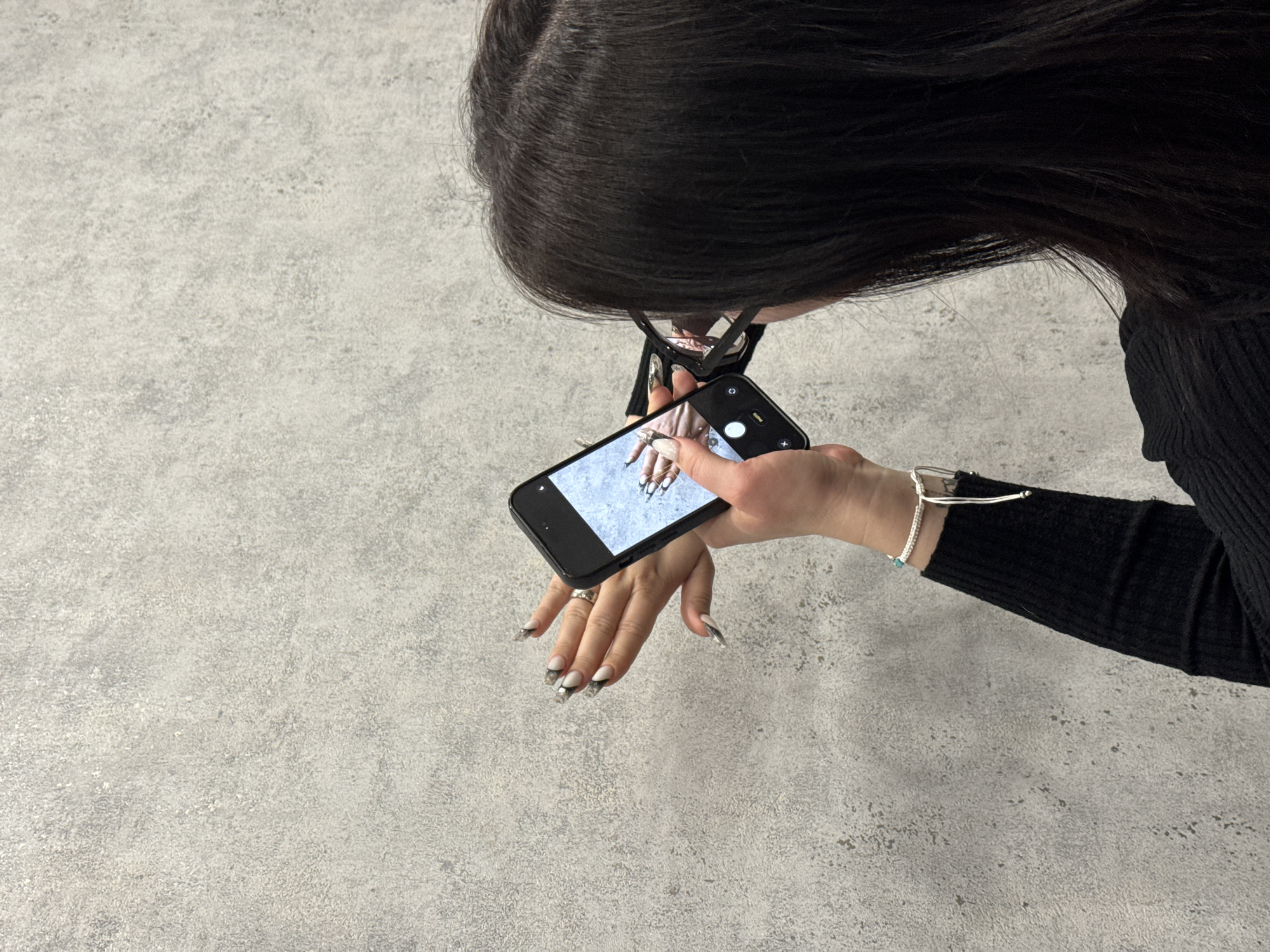 An overhead shot captures a person, likely female, with long, dark hair, wearing black-framed glasses and a black ribbed long-sleeved top. The person is holding a black smartphone in their right hand, pointing it downwards towards their outstretched left hand.
The activity clearly shows the person taking a picture or video of their own left hand, which is positioned in front of them. The screen of the smartphone displays an image of fingers with long, elaborately designed nails, mirroring the appearance of the hand being photographed. The nails feature a distinctive French tip style with black and silver or metallic grey accents. A silver-toned ring is visible on the ring finger of the left hand. On the person's left wrist, a delicate silver-colored bracelet adorned with a small turquoise bead is visible.
The background consists of a uniform, light grey textured surface, resembling concrete or a speckled stone floor or tabletop. The lighting appears even, suggesting an indoor setting. The composition focuses heavily on the action of self-photography, highlighting the intricate manicure. No specific time of day or location can be determined, and no discernible text is visible on the phone screen or elsewhere in the image.