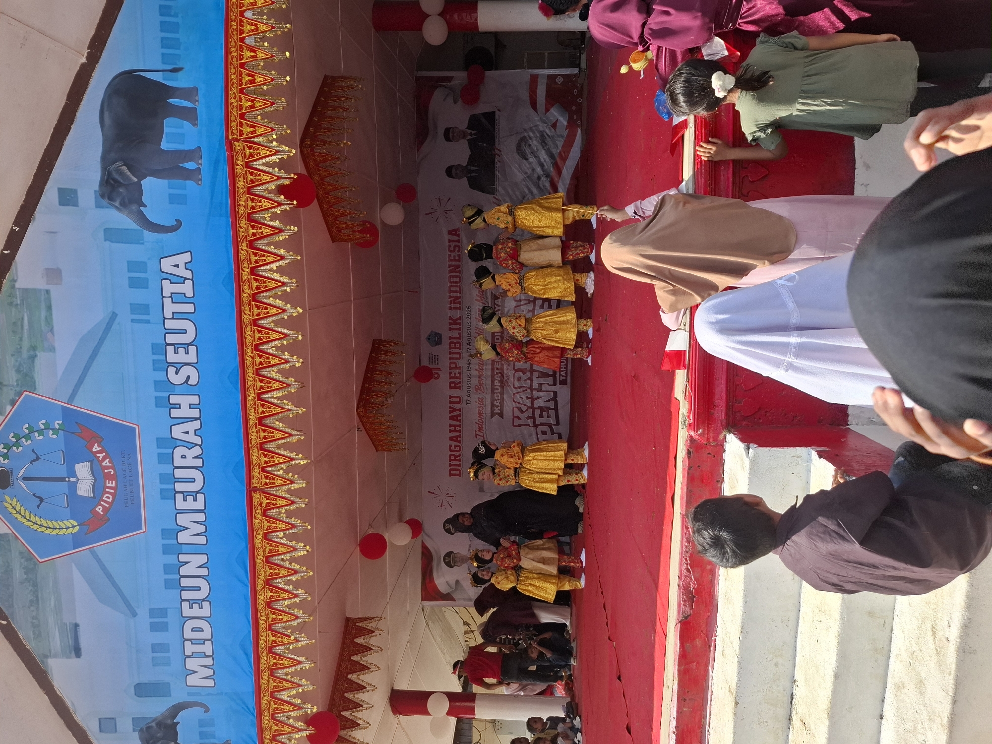 On a sunny day, a performance is taking place on an outdoor stage at Rhieng Blang, Indonesia. The stage is covered with a red carpet and decorated with red and white banners and balloons. A large banner at the back of the stage features the text "MIDEUN MEURAH SEUTIA" and an image of an elephant. Below this, another banner proclaims "DIRGAHAYU REPUBLIK INDONESIA". A group of young girls, dressed in traditional yellow and gold attire, are performing on stage. Some adults are present on the stage, including women in black and burgundy clothing and a man in a red shirt. In the foreground, several children are seated on white steps, watching the performance. The overall atmosphere appears festive and celebratory.