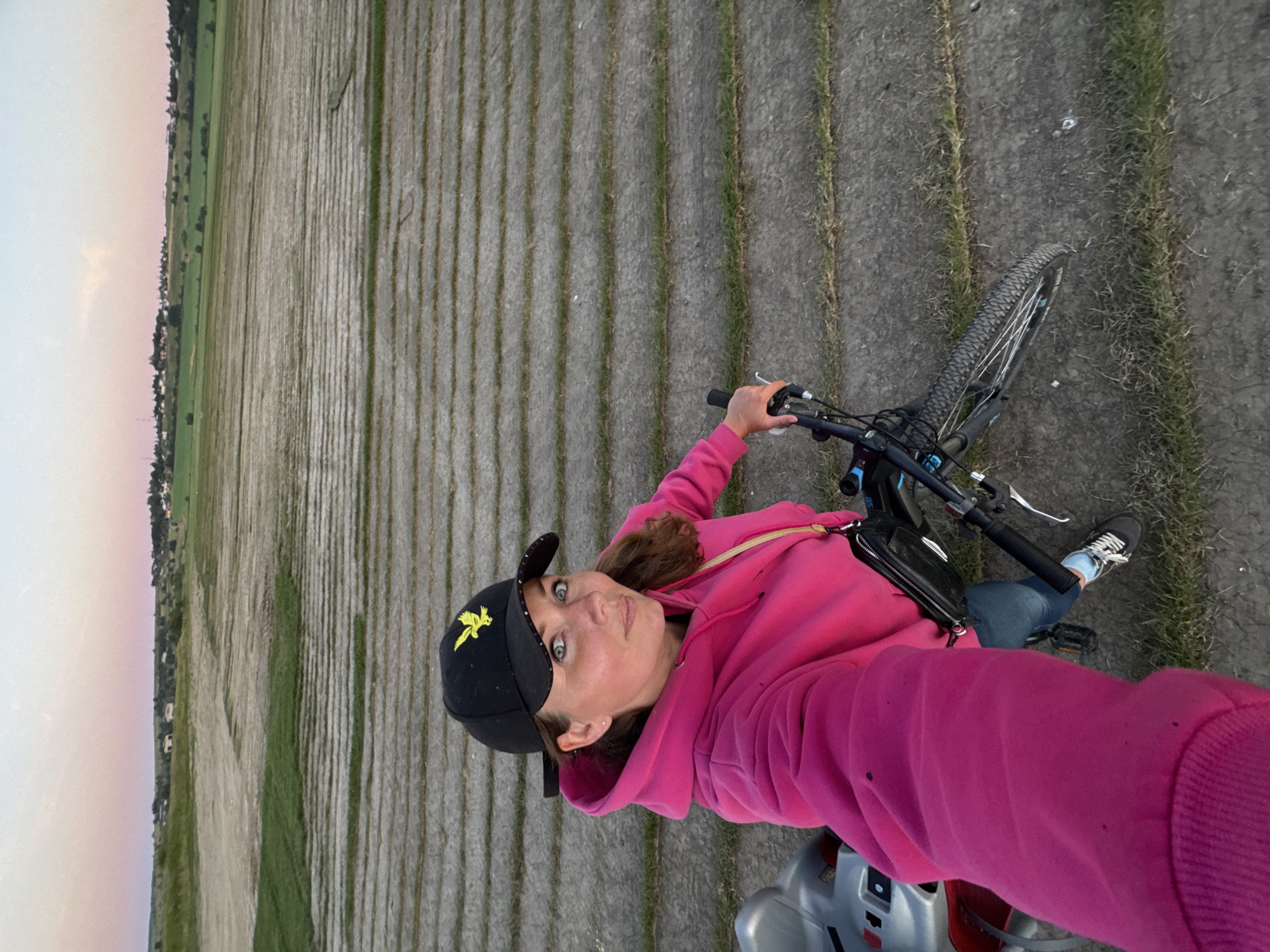 A woman wearing a pink hoodie and a black baseball cap with a yellow bird emblem is taking a selfie with her bicycle. She is standing on a paved surface with grassy strips. In the background, there is a vast agricultural field with visible rows of crops and a distant town with buildings under a soft pink and blue sky. The scene appears to be outdoors in Kempelevo, Russia, possibly during dusk or dawn.