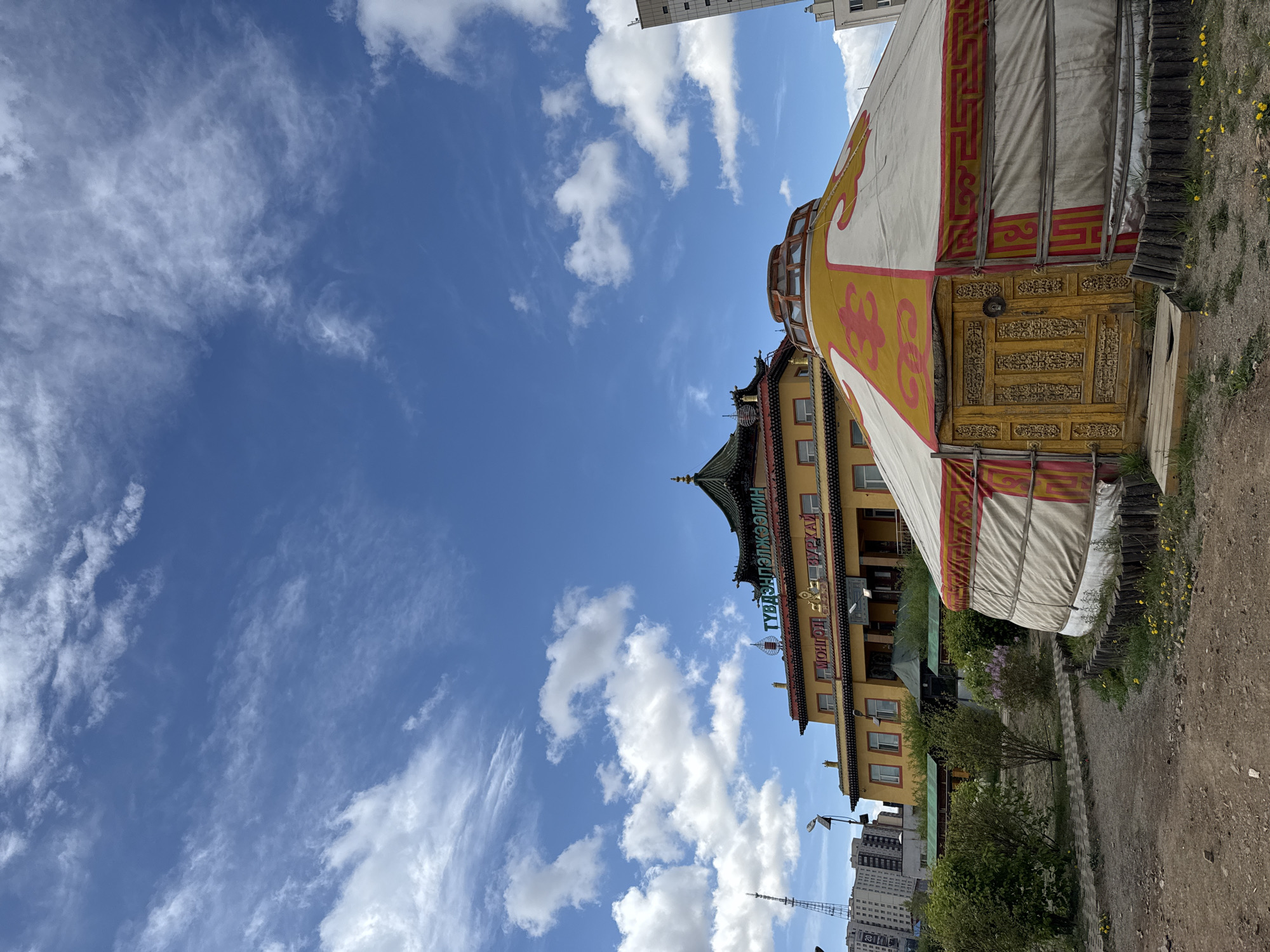 The image shows the Gandantegchinlen Monastery in Ulaanbaatar, Mongolia, with a traditional yurt (ger) in the foreground. The monastery building is multi-tiered with a green roof and ornate yellow facade. A blue sign with white lettering in Mongolian Cyrillic is vertically displayed on the building. The yurt is white with yellow and red traditional patterns. The sky is blue with scattered white clouds. Modern buildings can be seen in the background. The ground appears to be a dirt path with some vegetation.