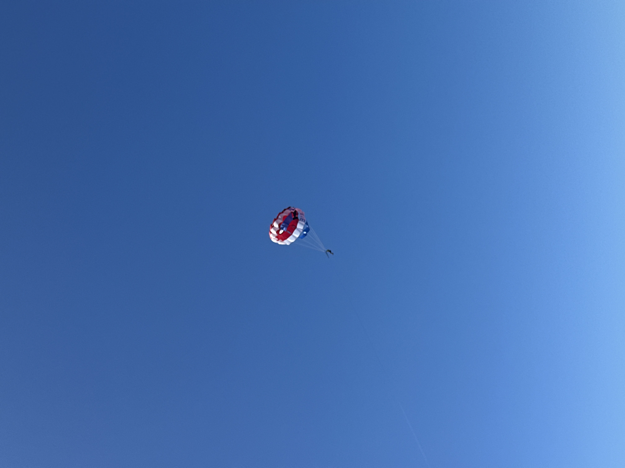The image displays a clear, vibrant blue sky dominating the frame, with a single parasailer as the central subject. A colorful parasail, featuring distinct red, white, and blue sections, is prominently visible in the upper-middle portion of the sky. Directly beneath the parasail, a small, dark, silhouetted figure of a person can be seen, suspended by lines.
The activity depicted is parasailing, with the individual soaring high above. The weather appears to be excellent, characterized by a completely cloudless and bright sky, indicative of a sunny day suitable for such aerial recreation. The blue of the sky shows a subtle gradient, appearing slightly deeper towards the top and left, and marginally lighter towards the bottom right.
A faint, thin line is visible extending downwards from the parasailer, presumably connecting to a tow boat that is out of frame. The focus is entirely on the parasailer and the vast expanse of the sky, conveying a sense of height and freedom.
There is no visible text within the image. Given the location context of Budva, Montenegro, this scene likely takes place over the Adriatic Sea, a popular setting for watersports and tourist activities like parasailing.