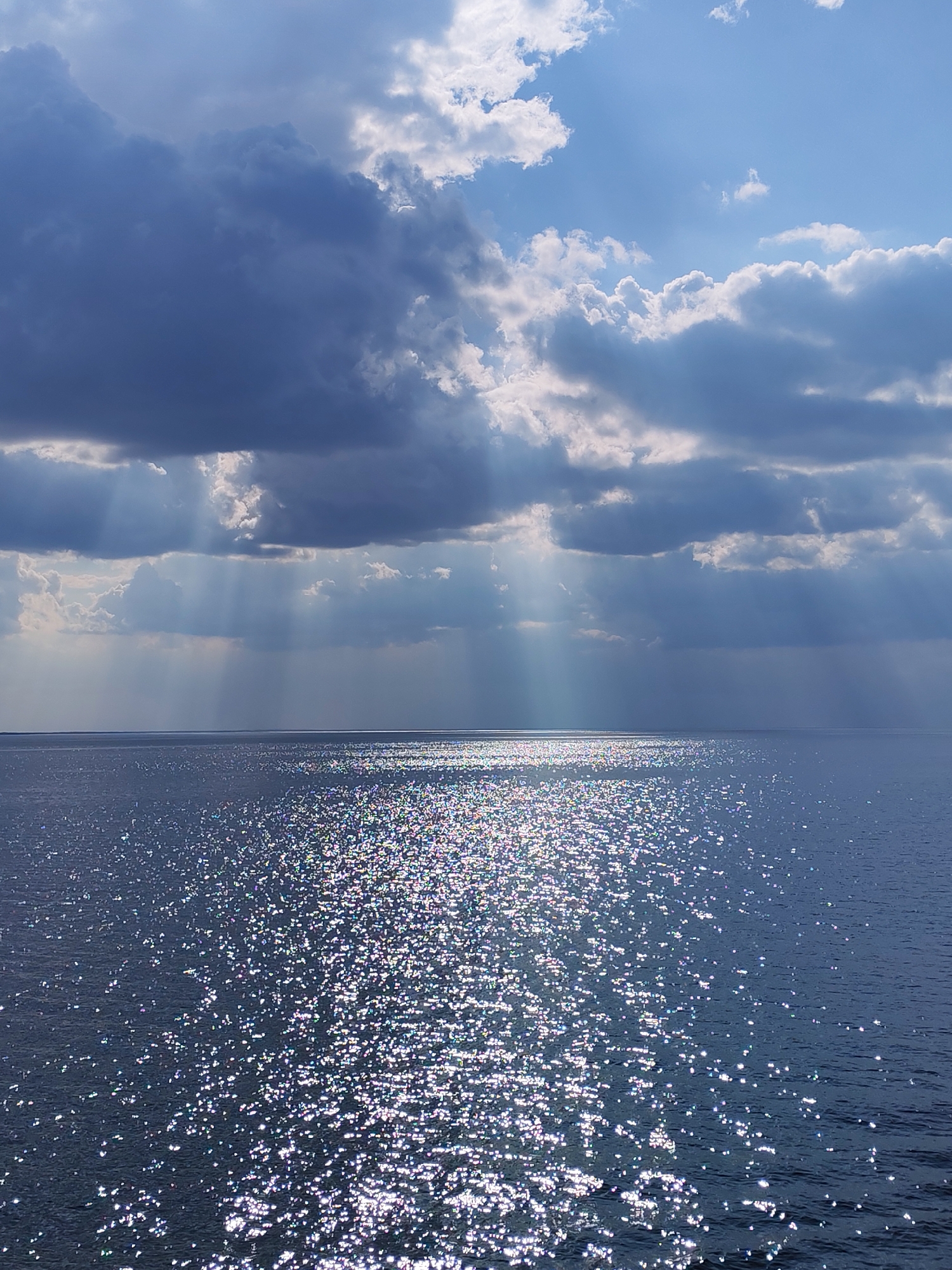 The image shows a body of water, possibly a lake or sea, under a sky with a mix of dark, imposing clouds and patches of bright blue sky. Sunlight streams down from behind the clouds in distinct rays, creating a dramatic and ethereal effect. The water's surface is covered in a shimmering, speckled reflection of the sunlight, creating a sparkling appearance. The horizon line appears distant and relatively flat. Given the location of Belozersk, Russia, it's probable the body of water is Lake Beloye.