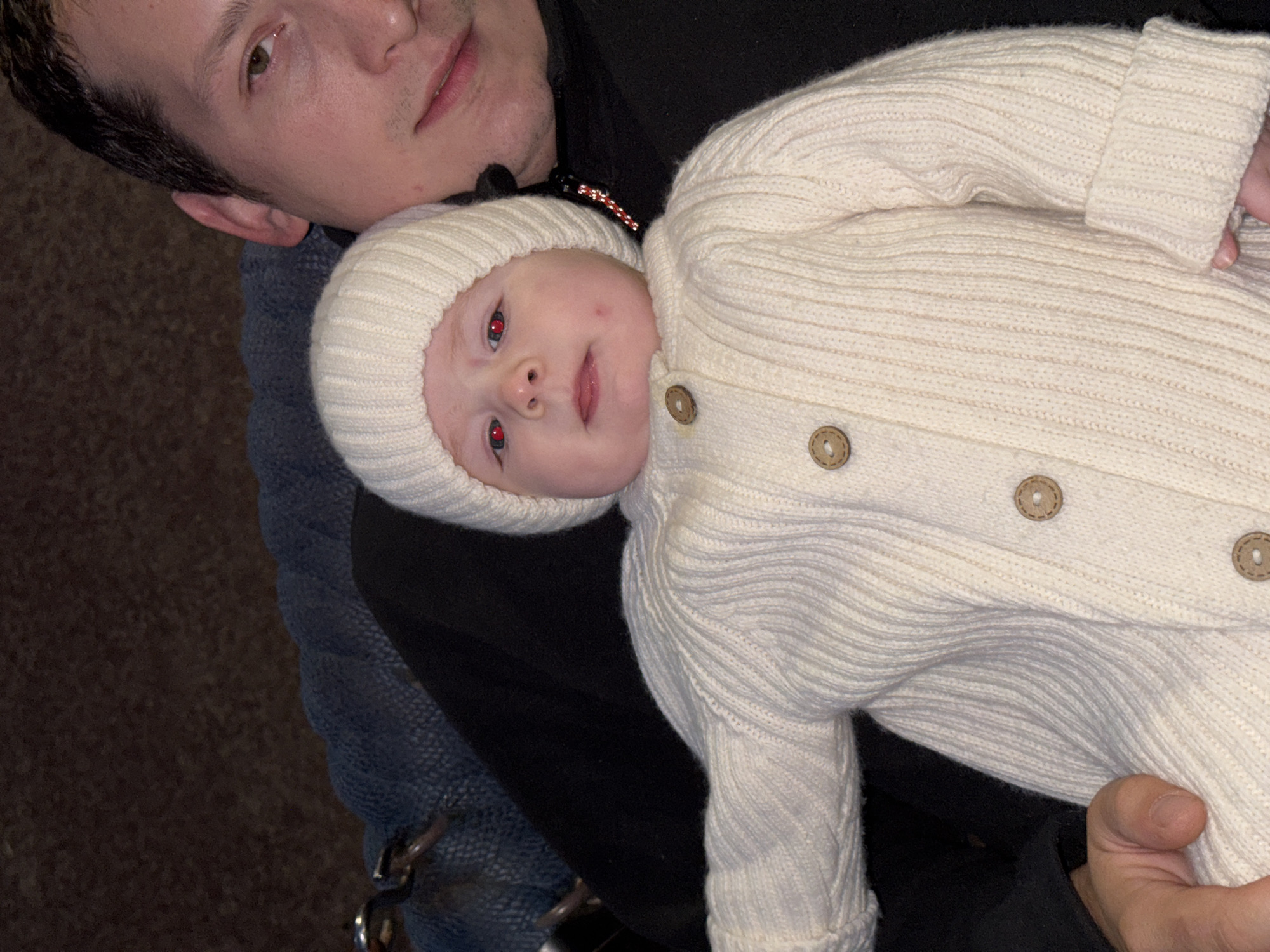 The image shows a close-up of a baby being held by a man. The baby is wearing a white knitted hat and a cream-colored knitted cardigan with wooden buttons. The baby has red-toned pupils. The man, whose face is partially visible, is wearing a black jacket. They are likely in a home environment, possibly a living room, indicated by the blurred background of what appears to be furniture or bedding. There is no information in the image that allows for the determination of the specific location within Noviy, Russia.