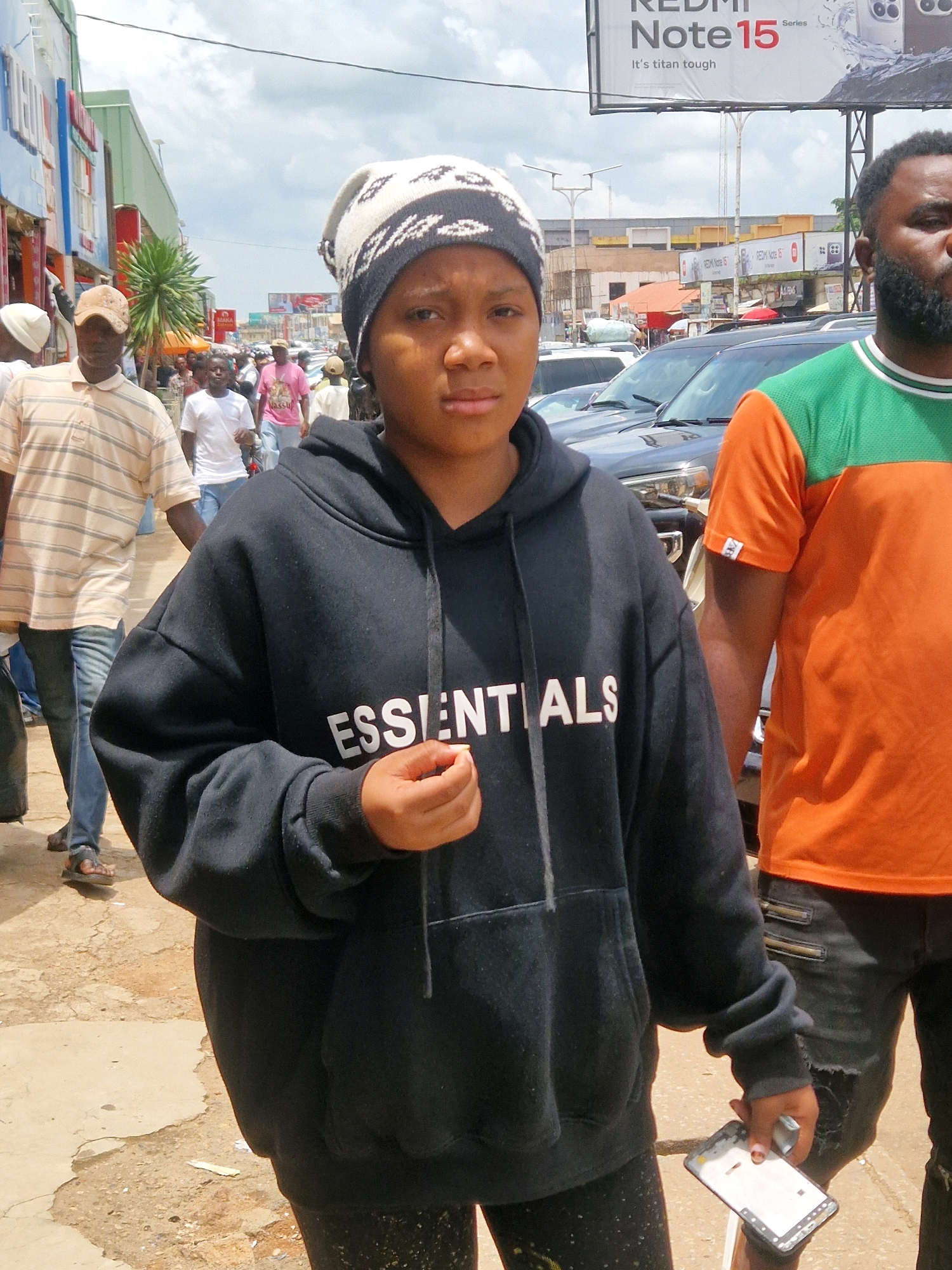 In Jos, Nigeria, on a cloudy day, a young person wearing a black hoodie with "ESSENTIALS" printed in white lettering on the front stands in the foreground. They also wear a black and white patterned beanie and dark leggings. Their right hand is held up, and they are holding a disassembled smartphone. To the right of the young person, an adult male with a beard wears a bright orange and green t-shirt and dark pants with zippered pockets. Behind them, several cars are parked on a street lined with shops. People are visible in the background, and a large billboard advertising "REDMI Note 15 Series" is prominent in the upper part of the image. The ground is a mixture of dirt and cracked concrete.