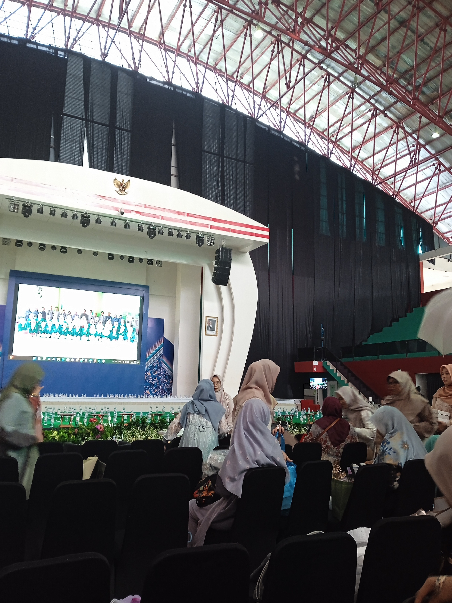 The image shows an auditorium-like setting with a stage, screen displaying a group photo, and rows of black chairs in the foreground. Several women wearing hijabs are scattered around the seating area, some standing and moving. The stage is decorated with flowers and water bottles. The structure of the building features a red and white arched metal frame ceiling, with black curtains covering a portion of the background. A coat of arms is visible on the stage, indicating a possible government or official event. The location is Unknown, Unknown.