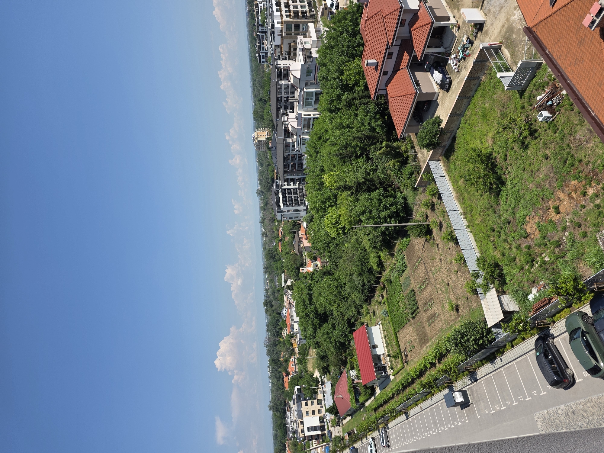 This is an aerial, high-angle, full shot of a residential neighborhood on a bright, sunny day. The scene is located in the Plovdiv, Bulgaria area, specifically in the western part of the city. The sky is a clear, bright blue with a few scattered, fluffy white clouds. The foreground is dominated by a paved parking lot with clearly marked parking spaces, some of which are occupied by vehicles. A dark gray minivan and a dark green SUV are parked in the lot. Adjacent to the parking lot is a patch of green grass and some landscaping.
Further into the image, there are several residential buildings. The closest buildings are single-family homes with red tile roofs. Some of these homes have garages, with cars parked inside. One of these homes has a red metal roof. Beyond these, there are more houses, interspersed with dense green trees. In the mid-ground, there is a cultivated garden area with rows of plants. Further back, several modern apartment buildings with flat roofs and light-colored facades are visible, surrounded by more greenery. The background shows a distant landscape with trees and possibly mountains, suggesting the outskirts of the city. The overall impression is one of a well-developed, suburban area with a mix of housing types and ample green space.