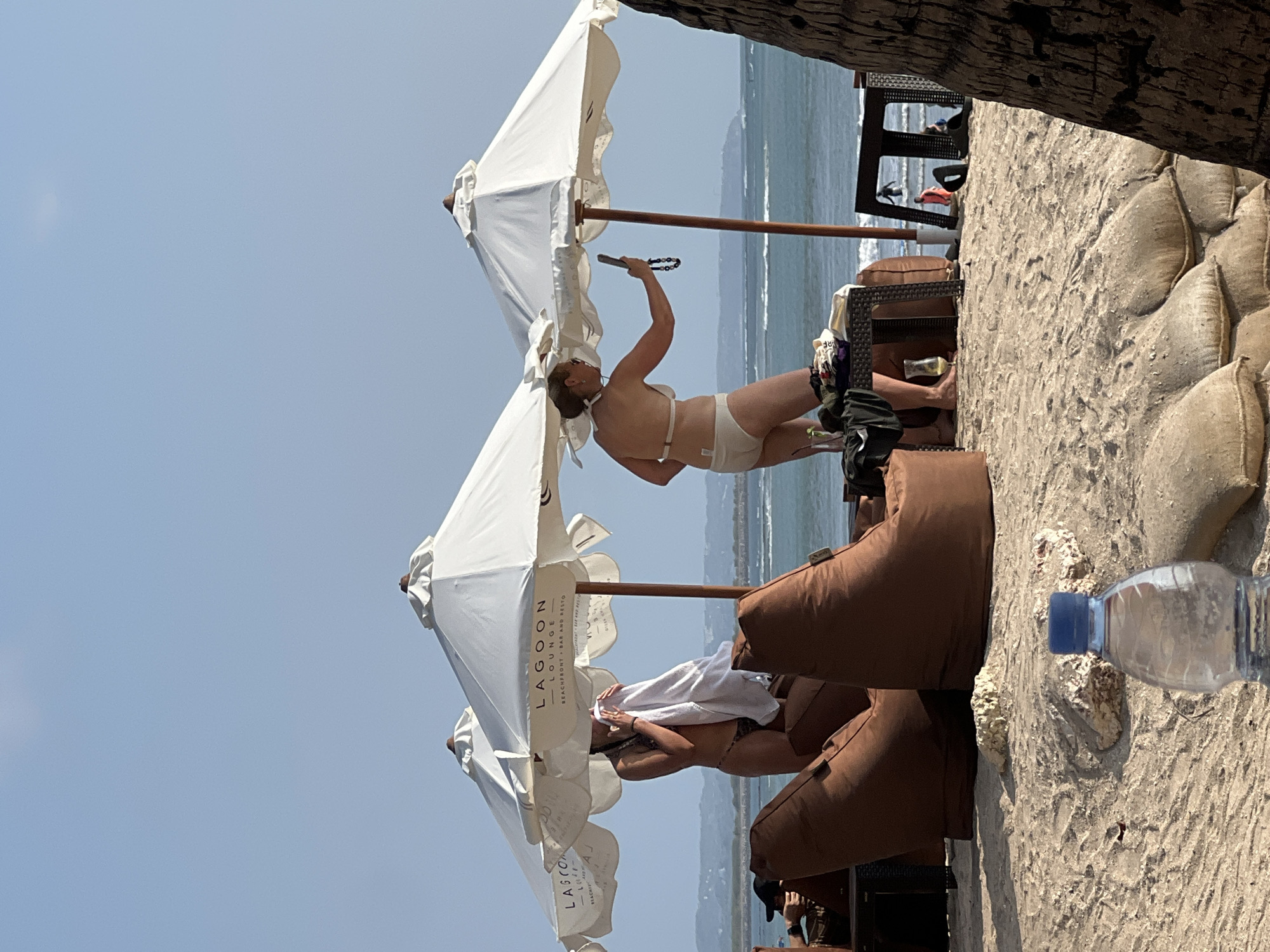 On a bright, sunny day at the beach in Kuta, Indonesia, two women are seen relaxing under large white umbrellas. The woman on the left, wearing a patterned bikini, is drying herself with a white towel. The woman on the right, in a white bikini, is holding a phone and appears to be taking a selfie. Both women are positioned on a sandy beach, with brown beanbag chairs and a small table with drinks nearby. In the background, the ocean stretches out to the horizon, with gentle waves and a few distant figures in the water. The umbrellas are branded with "Lagoon Lounge," suggesting a beach club or restaurant. The overall scene depicts a leisurely day at a popular tourist destination.