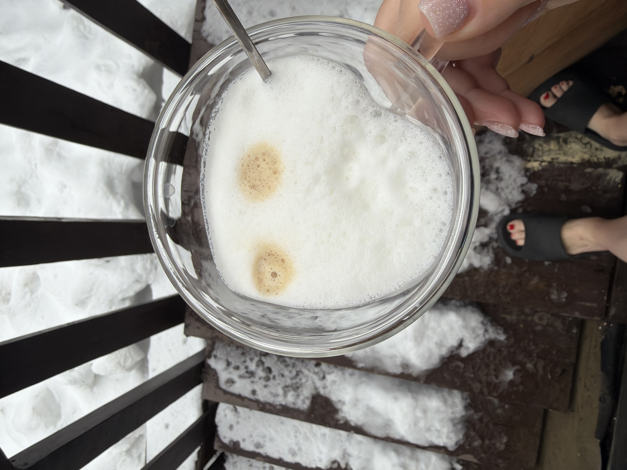 Here is a description of the image:
The photograph shows a top-down view of a glass mug filled with a foamy beverage, likely a latte or cappuccino, held by a person with manicured nails. A spoon is partially visible within the mug. The mug sits above a wooden staircase covered in snow. The staircase appears to be outdoors, as evidenced by the presence of snow. Black railing is visible to the left of the stairs. Two feet clad in black sandals are also visible near the upper-right quadrant of the photograph. The photograph was taken in the city of Chaikovsky, Russia.