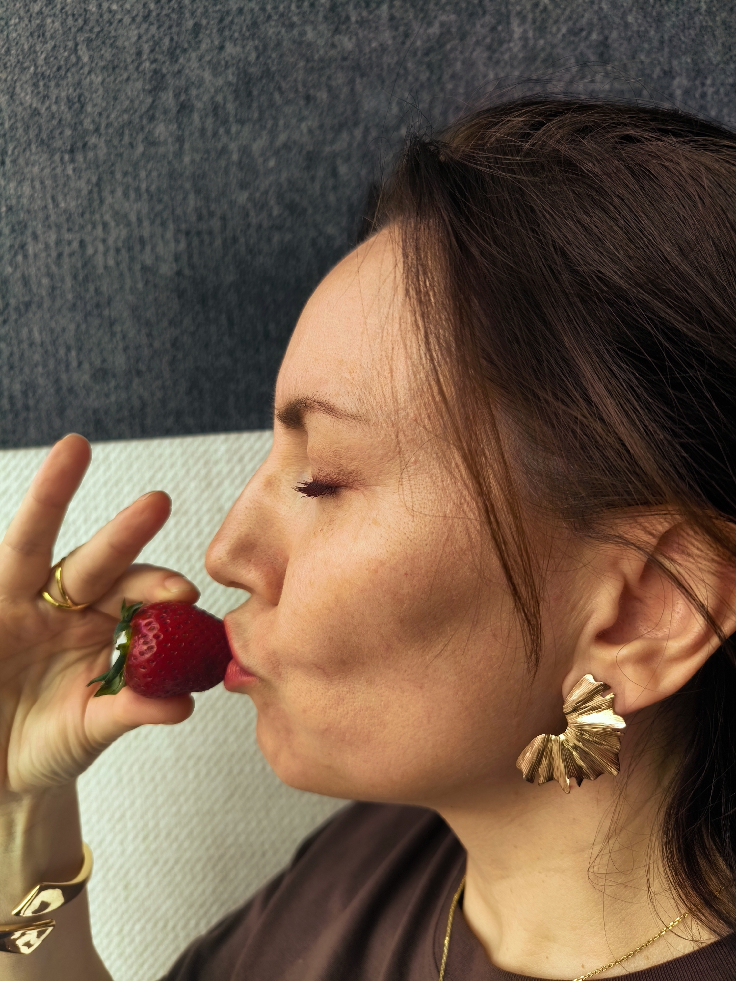 A woman is photographed in profile, positioned near what appears to be a white wall or cushion on one side, and a gray textured background on the other. Her eyes are closed, and she is holding a large strawberry up to her lips as if about to take a bite. She wears gold jewelry, including a ring on her finger, a cuff bracelet on her wrist, and prominent decorative earrings. Her hair is dark and styled around her face. She is wearing a dark-colored shirt. The location of the scene is Unknown, Unknown.