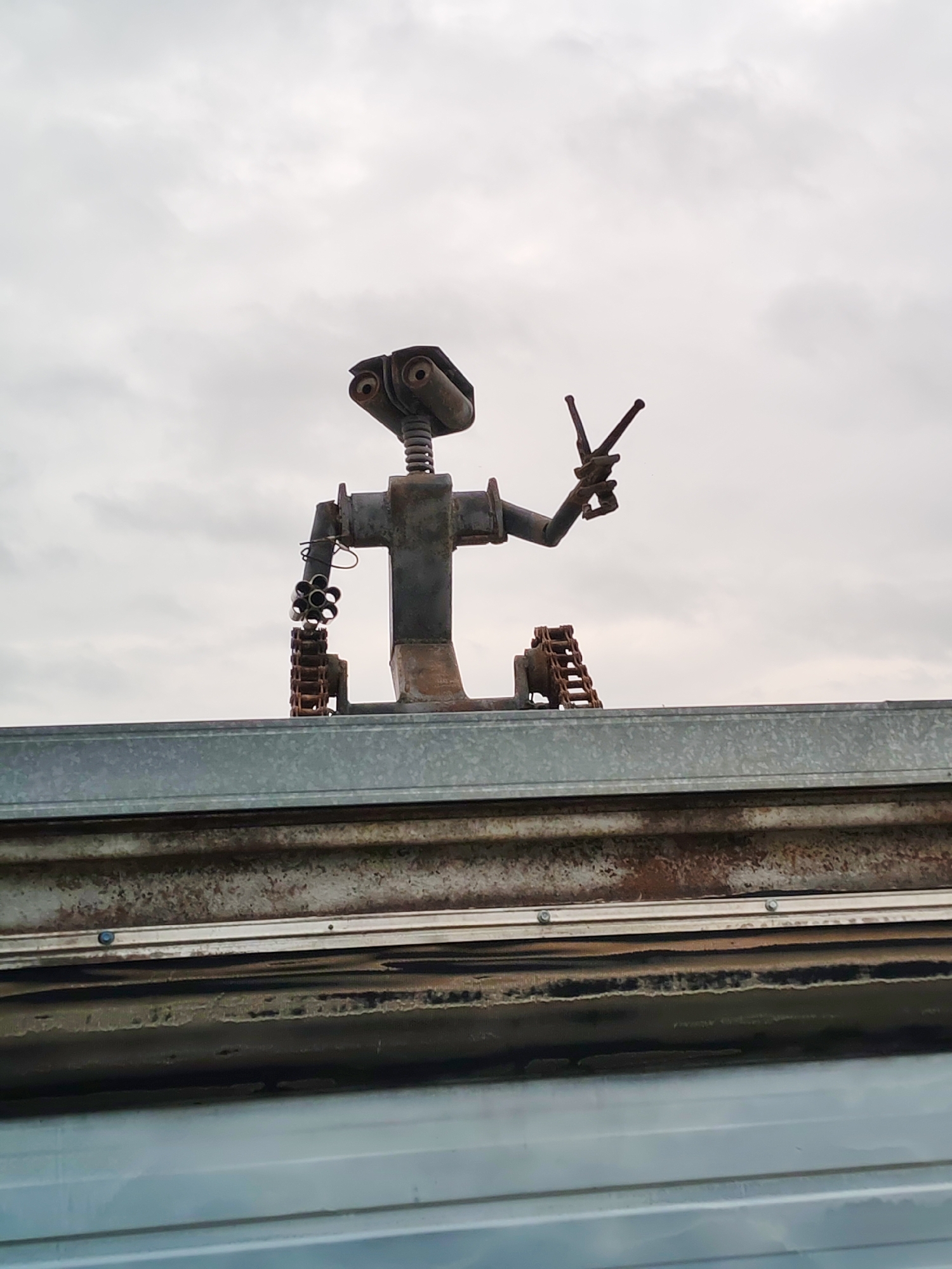 A close-up, low-angle shot captures a metallic robot sculpture in front of a cloudy sky. The robot, constructed from various metal parts, is positioned on top of a corrugated metal roof. It has two round, lens-like eyes, a spring-like neck, and two arms. One arm is bent at the elbow and features a circular attachment with multiple tubes, resembling a Gatling gun. The other arm is raised and bent, with its hand formed into a peace sign. The robot's body is a metal structure with visible bolts and joints. The foreground is dominated by the textured, weathered surface of the corrugated roof, which shows signs of rust and age. Below the robot, there are two horizontal bands of metal, one lighter gray and the other darker, likely part of a larger structure. The background is a uniformly gray, overcast sky with faint cloud formations. The overall scene appears to be outdoors, possibly in an industrial or artistic setting.