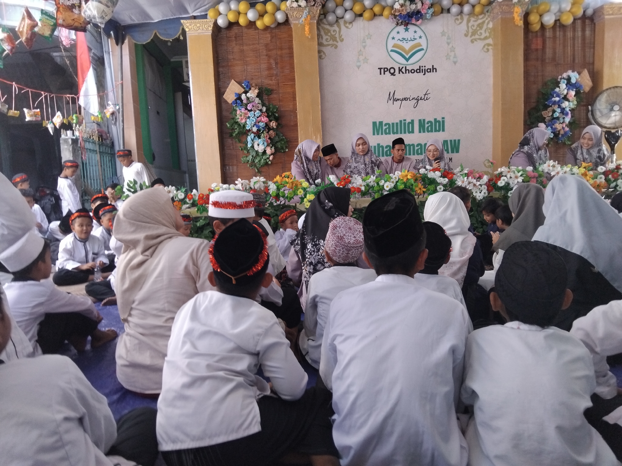 The image depicts an indoor gathering, likely a religious event, held under a tent structure. The setting appears to be in Kepuh Kiriman, Indonesia, as indicated by textual elements in the background. The event is themed around "Maulid Nabi," a celebration of the Prophet Muhammad's birthday.
In the foreground, several children are seated on a blue mat, facing away from the camera. They are dressed in white attire and many are wearing peci (traditional caps). Some children have red and gold decorative bands on their caps.
The background features a stage area with a banner that reads "TPQ Khodijah" and "Memperingati Maulid Nabi Muhammad SAW". A variety of floral arrangements adorn the stage. Several adults, predominantly women wearing hijabs, are seated behind the floral arrangements. Some of them are engaged in conversation or looking towards the audience.
To the left of the image, there is a partial view of a red and white Indonesian flag hanging vertically. Further to the left, a fence and some decorative elements suggest an outdoor component or entrance to the venue. The overall atmosphere seems to be one of communal celebration and religious observance.