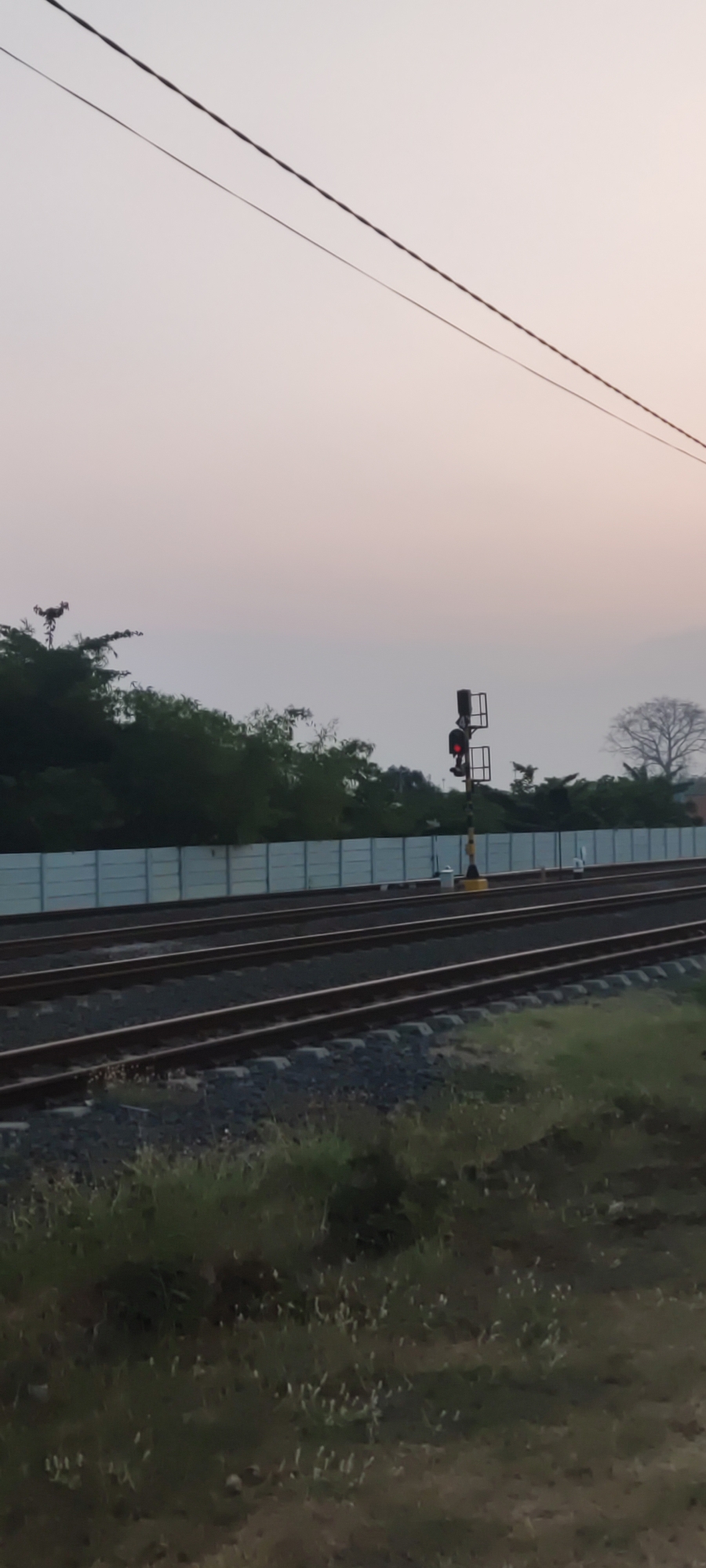 The image depicts a railway scene at dusk in Babakan, Indonesia. Twin parallel railway tracks stretch across the frame, bordered by gravel and sparse vegetation. A light blue concrete wall runs along the left side, separating the tracks from a dense line of green trees. A solitary railway signal, displaying a red light, stands prominently on the right side, its pole marked with yellow and black stripes. Above the scene, two dark wires are strung across the pale, hazy sky, suggesting the presence of overhead electrical lines. The overall impression is one of a quiet, somewhat desolate industrial landscape under a soft, fading light.