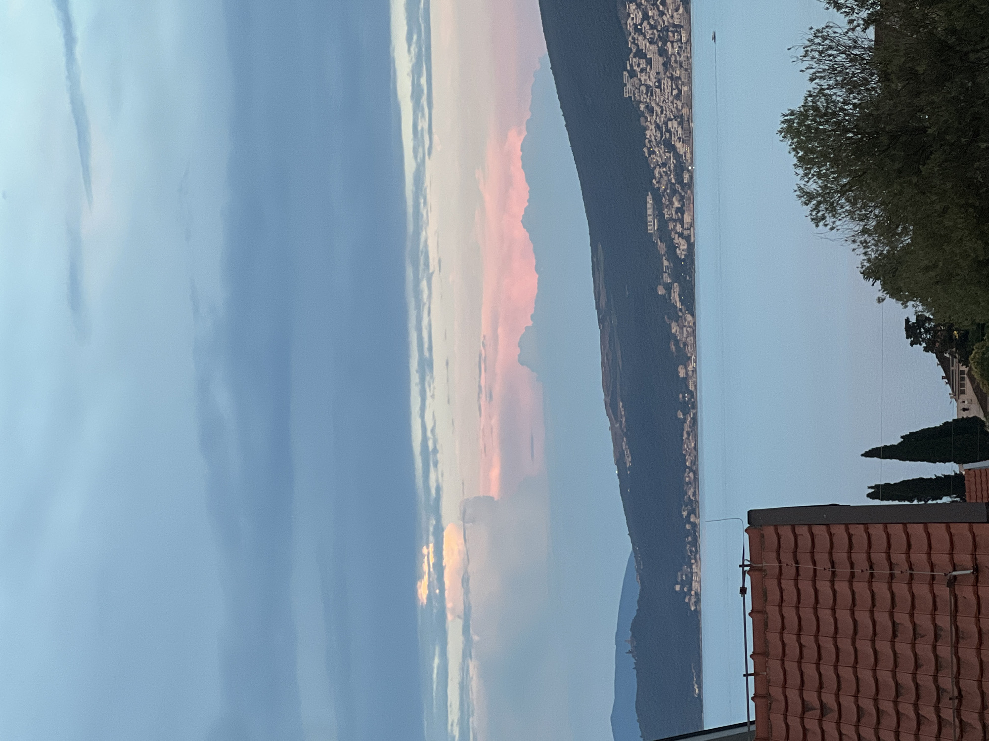 This is a vertically oriented, slightly low-angle photograph of a coastal scene at dusk in Baošići, Montenegro. The dominant subjects are the sky, a body of water, and a hillside town. The sky exhibits a gradient of soft blues and pastel pinks, suggesting the final moments of sunset or the early stages of twilight. Wispy clouds are scattered across the sky, catching the fading light and adding texture.
The body of water, likely the Adriatic Sea, is calm and reflects the muted colors of the sky. On the far side of the water, a dark, forested hillside rises steeply, densely populated with buildings forming a town that stretches along the coast. The lights of the town are visible, twinkling like scattered jewels against the darkening landscape.
In the foreground, a red-tiled roof of a building juts into the frame from the bottom right, providing a sense of immediate perspective and human habitation. To the right of the roof, the dark green foliage of a tree partially obscures the view, and further back, two tall, conical cypress trees stand sentinel.
The overall mood is tranquil and serene, evoking a sense of peaceful evening by the sea. The time of day is clearly dusk, characterized by the soft, diffused light and the transition from day to night. There is no visible text in the image.
