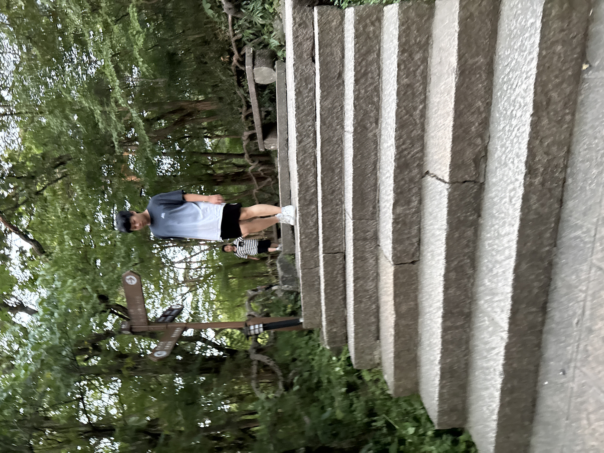 In the city of Huangshan District, China, a young man walks down a wide stone staircase, surrounded by lush green trees. He is wearing a dark t-shirt with a faded gradient and black shorts, paired with white sneakers. Behind him, further up the stairs, a child in a striped t-shirt and dark shorts is also walking. The staircase is made of large, rough-hewn stone blocks. To the left of the stairs, a signpost with multiple directional arrows stands among the trees, and a small lantern is attached to its base. A rustic wooden bench is visible in the background, nestled amongst the foliage. The environment appears to be a park or natural trail, with the dense greenery suggesting a temperate climate. The overall scene depicts a casual outing in an outdoor setting.