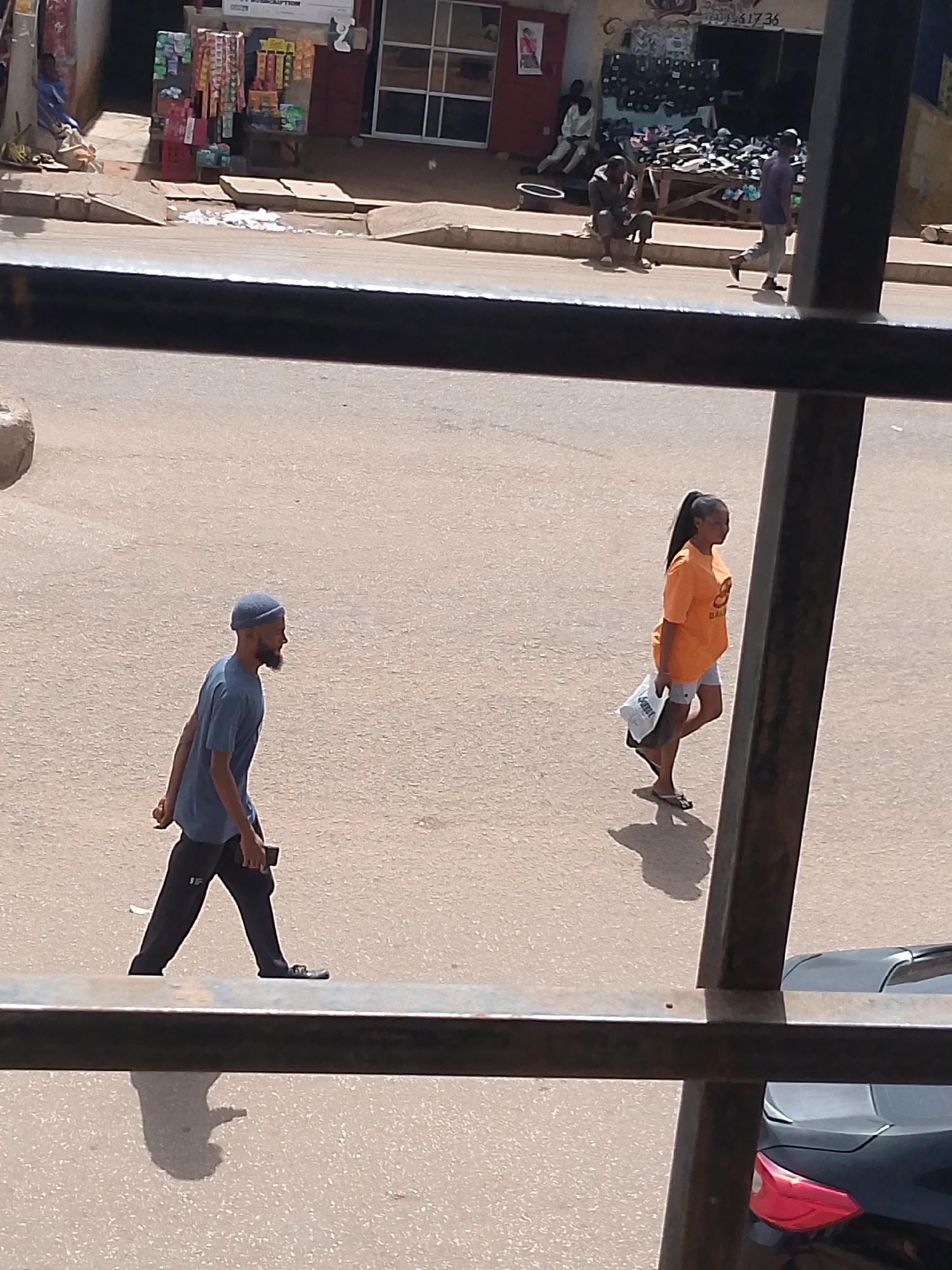 This is an eye-level, medium shot of a street scene in Bukuru, Nigeria, captured through a dark metal railing. In the foreground, a man wearing a blue t-shirt and a blue cap walks from left to right across a paved road. He has a beard and is holding a dark object in his right hand. Behind him and to the right, a woman wearing an orange t-shirt and light-colored shorts also walks from left to right. She has her hair in a ponytail and is carrying a white bag and a dark object in her left hand. Her shadow is cast on the road behind her. The background shows a dusty street with a few buildings. One building has shelves stocked with various items. There are other people visible in the background, some sitting and others walking. A black car with its taillight visible is parked on the right side of the road. The overall lighting suggests it is daytime, with strong sunlight casting shadows.