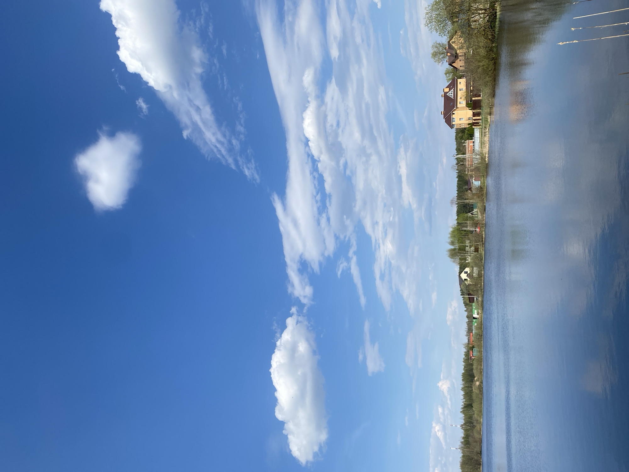 A vertical shot displays a lake and sky. The sky takes up most of the left side of the image, with cumulus clouds and a blue background. On the right side of the image is a lake. Houses line the bank of the lake, with trees and other greenery.