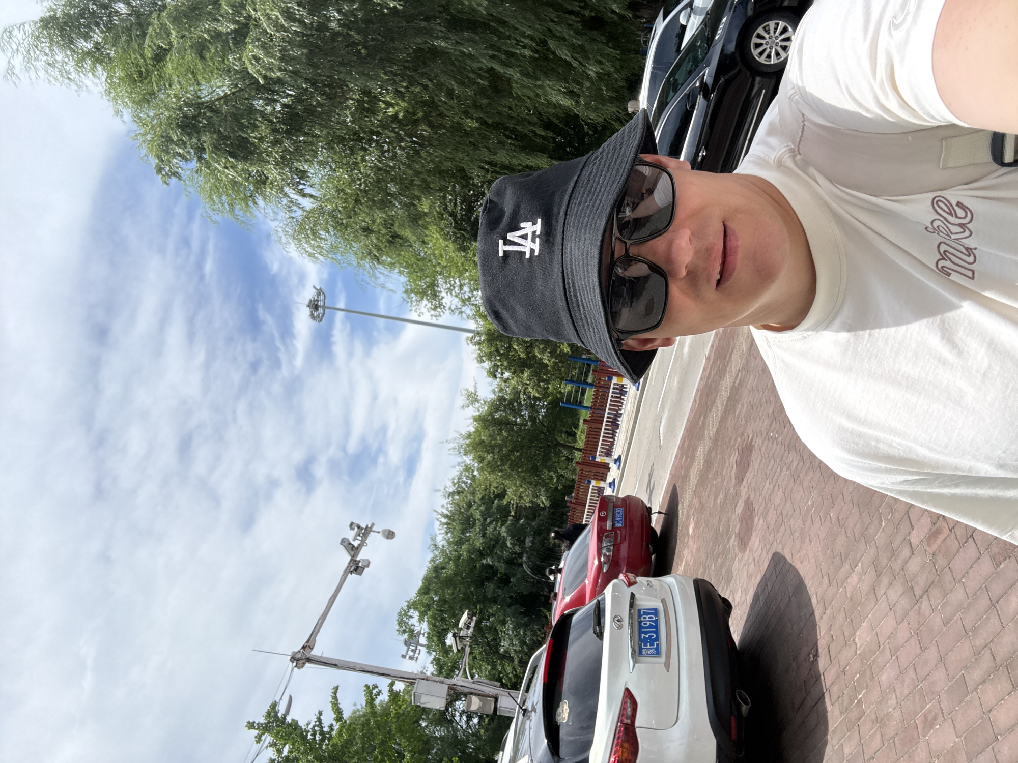 A man wearing a black bucket hat with the LA logo, sunglasses, and a white t-shirt is taking a selfie outdoors. He is standing on a red brick pathway. In the background, there are cars parked along a road, trees, a traffic light, and a blue sky with clouds. The license plate on the white SUV in the foreground reads "E 319B7". The red car behind it has the license plate "KC V9C30". The environment appears to be a street or parking area within the city of Qinhuangdao, China.
