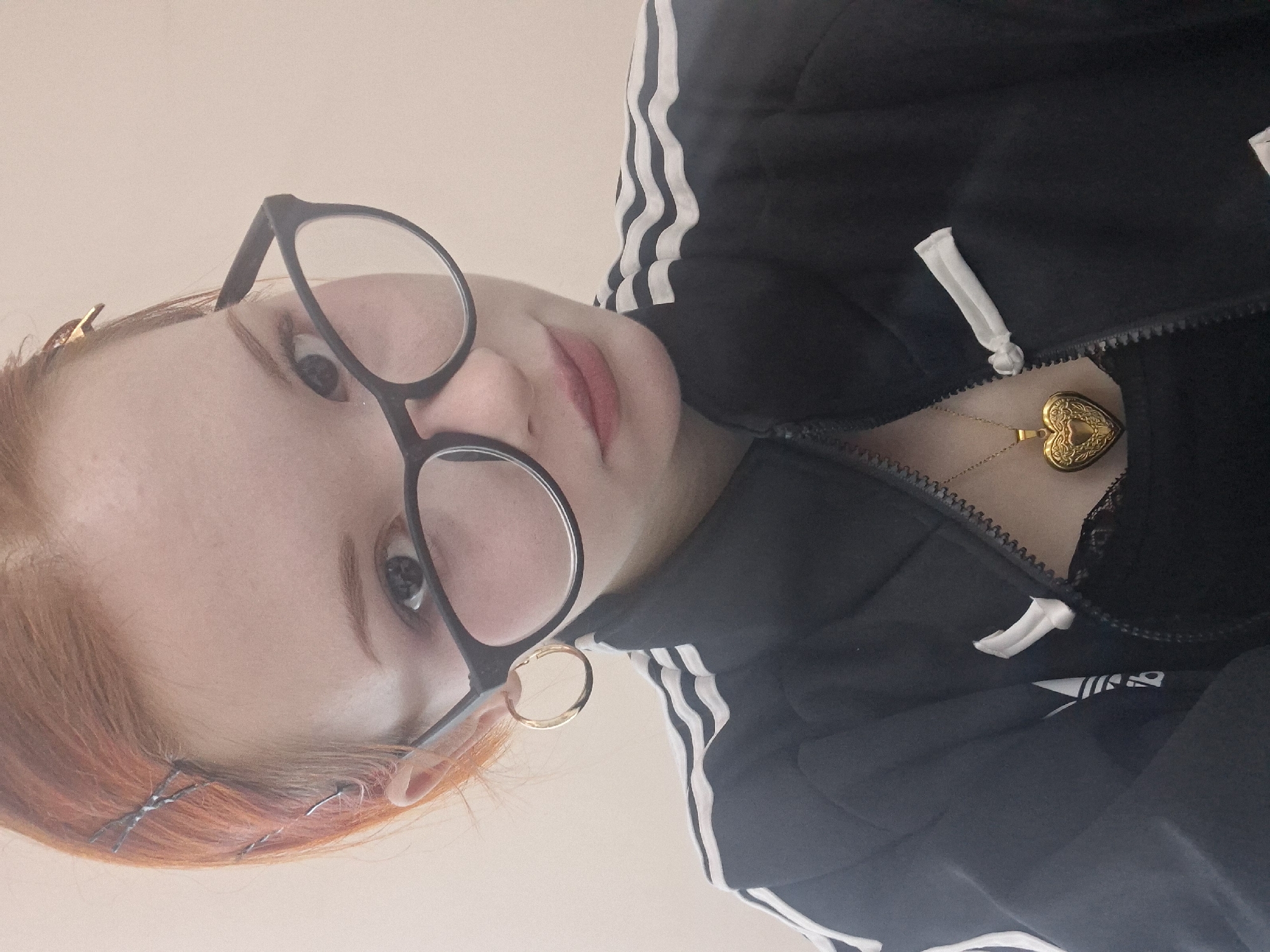 A close-up, tilted portrait captures a person with pale skin, reddish-orange hair secured with hairpins on the left side, and wearing black-framed glasses. The person is wearing a black Adidas track jacket with white stripes along the sleeves and a gold heart-shaped locket necklace. A gold hoop earring is visible on the left ear. The background is a plain, light peach color. The location in the city of Chaikovsky, Russia, is not discernable from the provided image.