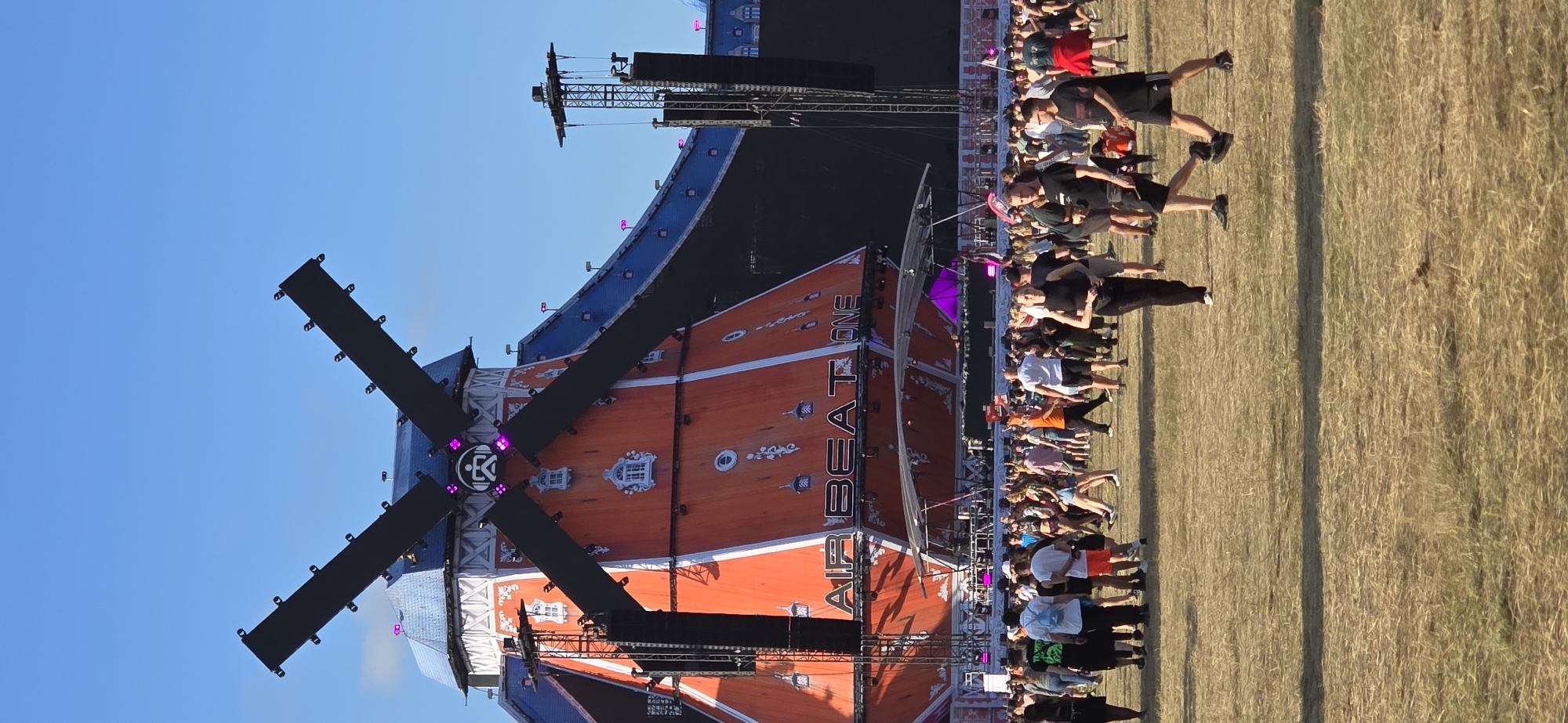 The image shows a crowd of people gathered in an outdoor field. In the background, a large, orange, windmill-shaped stage structure is visible, with the words "AIR BEAT ONE" prominently displayed on it. The windmill has black blades and is decorated with white window details. There are also large speaker towers on either side of the stage. The sky is clear blue. The people in the foreground are walking and standing, appearing to be attending an outdoor event or festival. The ground is covered in dry grass. The festival is the Airbeat One festival, which takes place annually in Neustadt-Glewe, Germany.