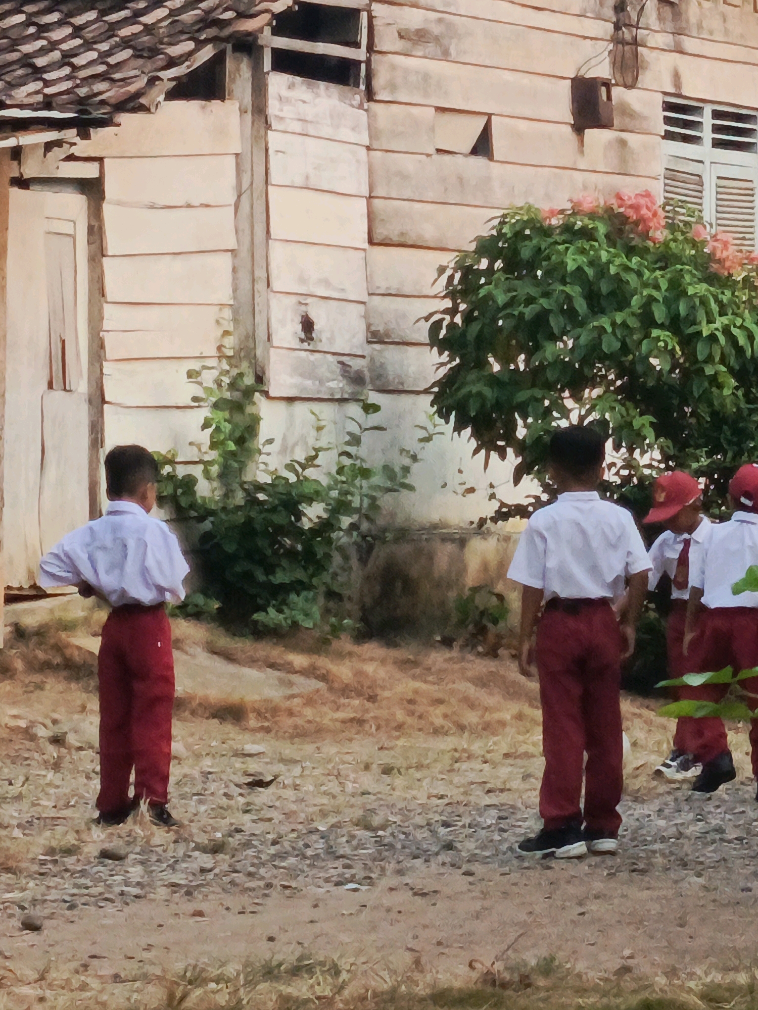 In front of a weathered, light-colored building with a tiled roof and wooden siding, four children in school uniforms stand on a dirt and gravel surface. The child on the far left, facing away from the camera, wears a white shirt and red pants, with hands on hips. To the right, three other children, also in white shirts and red pants, are visible from the back. The two children on the right are wearing red caps. One of them is slightly turned towards the left. Lush green foliage with pink flowers grows near the building. The scene appears to be an outdoor area, possibly a schoolyard, in Megang Sakti II, Indonesia, given the architectural style and vegetation.