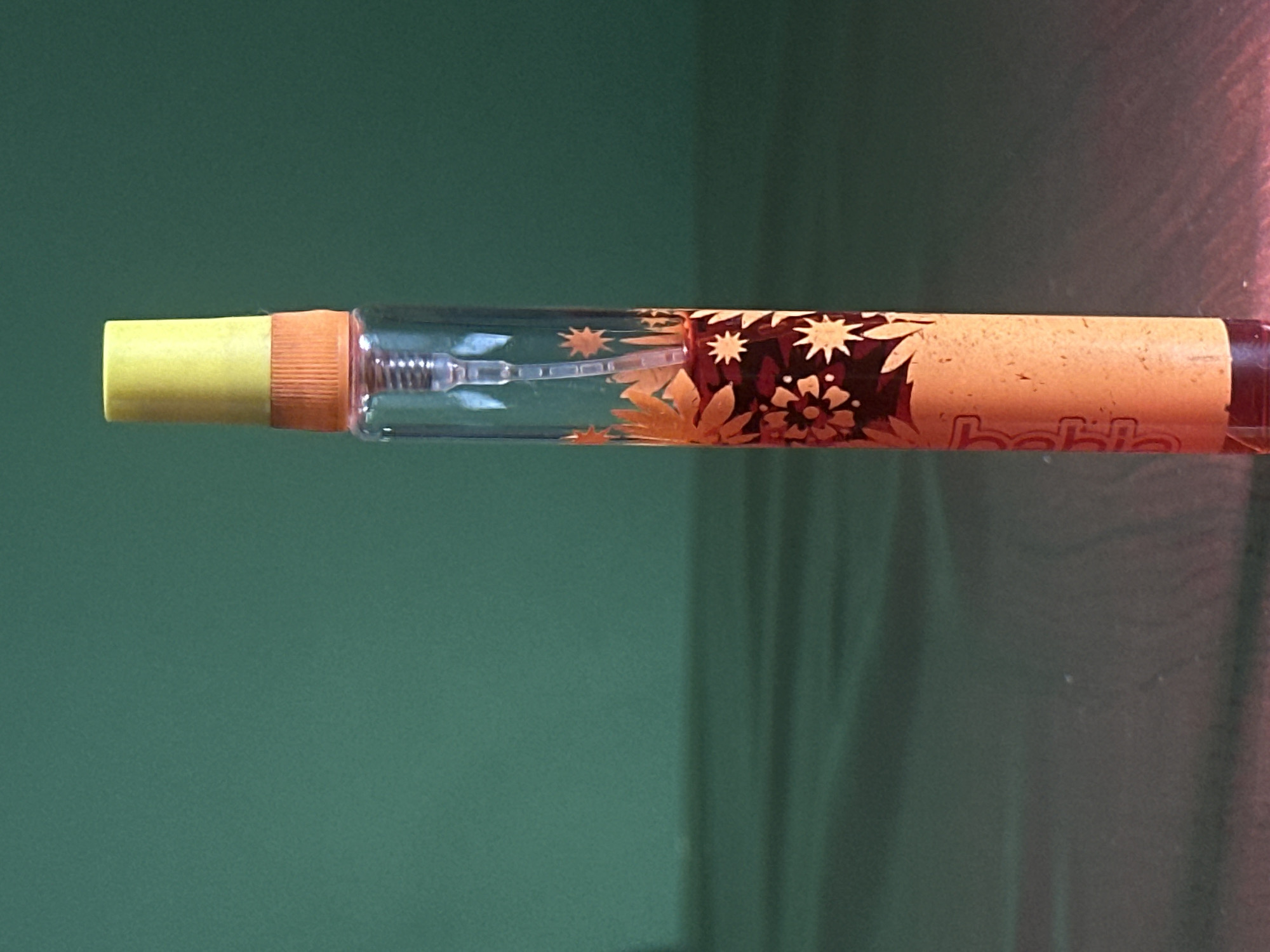 A clear, cylindrical glass perfume atomizer with an orange and yellow cap stands upright on a reflective dark surface. The bottle is decorated with an orange floral pattern on a dark background, and some of the liquid inside appears to be a dark red color. The background is a solid, dark green color, with subtle horizontal reflections visible on the surface the bottle is placed on. The lighting creates a soft shadow to the left of the bottle. There is no indication of the city of Lobacheve, Ukraine in the image.