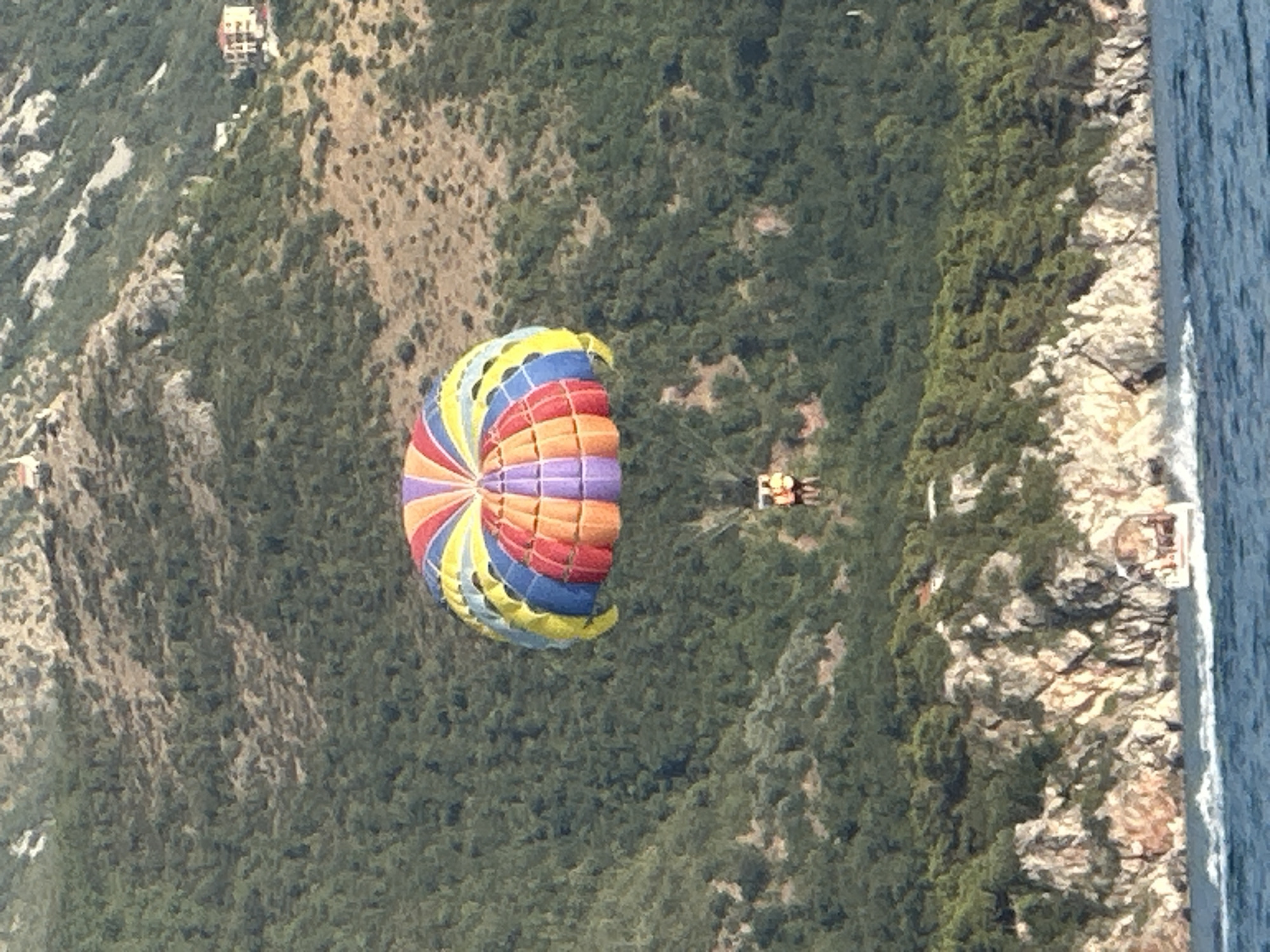 An outdoor, elevated, medium shot captures a vibrant, multicolored parasail with yellow, orange, red, blue, and purple sections floating against a backdrop of a densely forested, rocky mountainside. Two individuals are suspended in a harness beneath the parasail. In the lower portion of the image, a white boat with passengers navigates the blue water, leaving a foamy wake. The scene appears to be situated near Boreti, Montenegro, likely along the Adriatic coast, given the presence of rugged coastal terrain and the characteristic Mediterranean vegetation.