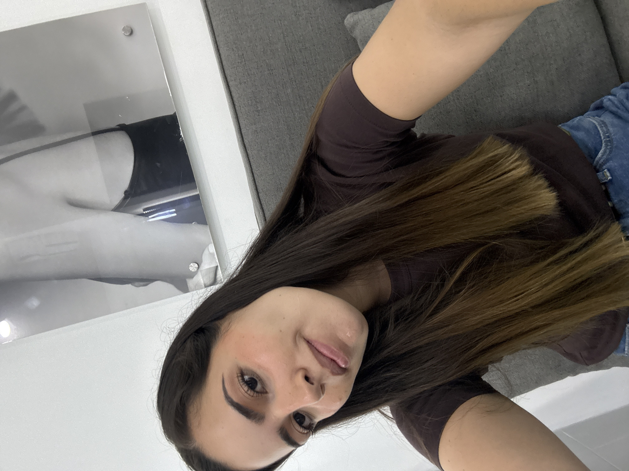 A woman with long, brown hair is posing for a selfie. She is wearing a brown top and blue jeans. She is sitting on a gray couch. Behind her is a framed picture. The location cannot be determined from the image.