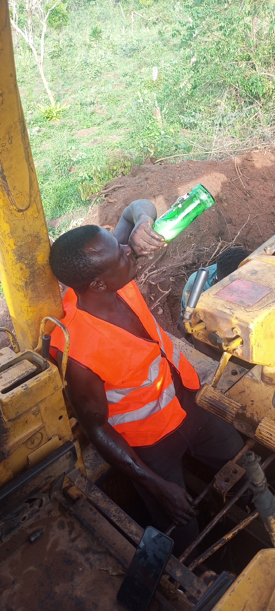 The image depicts a man taking a break while working with heavy machinery in Otunade, Nigeria. The primary subject is a dark-skinned man, seen from a high angle, seated within what appears to be the cabin or operator's area of a yellow excavator or bulldozer. He is wearing a bright orange safety vest with reflective grey stripes.
The man is actively drinking from a green glass bottle that contains a frothy, light-colored liquid, holding it to his mouth with his right hand. His skin, particularly on his arms and face, appears dirty or sweaty, indicative of physical labor. A black mobile phone with a dark screen lies on the dirty metal floor of the machinery, near his left leg.
Partially visible in the background, near the machinery's controls and within a dug-out area, is the head and shoulder of another person. The immediate surroundings consist of disturbed brown earth and exposed tree roots. Beyond the machinery and excavation, the background is filled with abundant green vegetation, including trees and dense foliage, characteristic of a natural, rural setting. The scene is brightly lit, suggesting it is daytime. No distinct text is visible on the bottle, machinery, or phone screen. The overall impression is of a worker resting during a task in an outdoor construction or excavation environment.