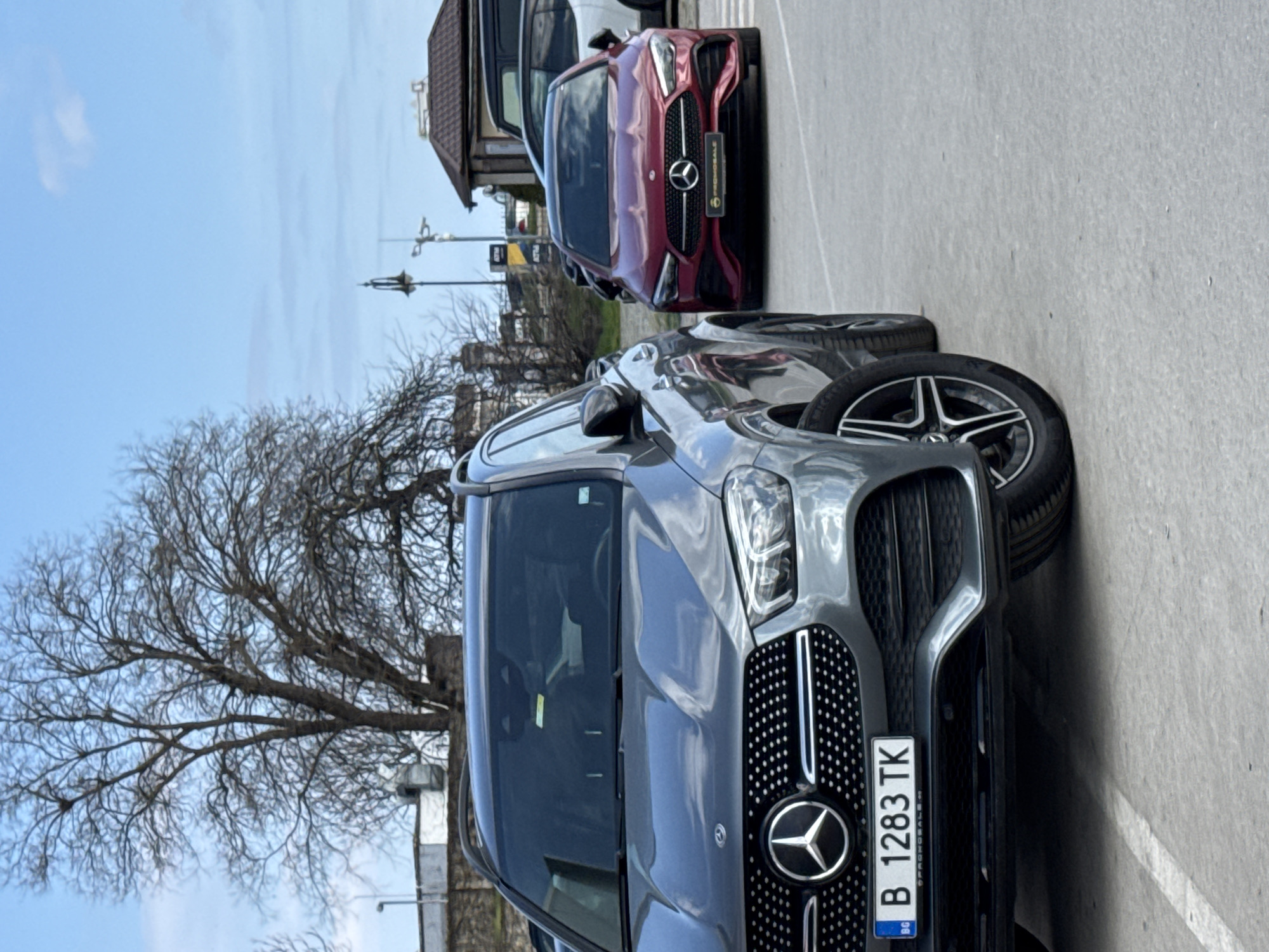 The image shows two Mercedes-Benz vehicles parked on a paved surface, likely a parking lot or street. The foreground vehicle is a gray Mercedes-Benz GLE SUV, with a Bulgarian license plate "B 1283 TK" visible. Behind the GLE is a red Mercedes-Benz coupe, also parked. The environment includes a bare tree on the left, buildings in the background with street lamps, and a partly cloudy sky. Given the Bulgarian license plate and architectural style, the location is presumed to be in Varna, Bulgaria. No people are visible. The image primarily captures static parked vehicles.