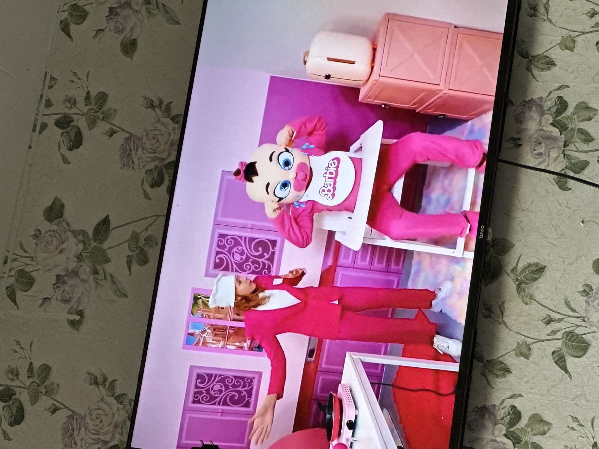 The image displays a television screen showing a vibrant, pink-themed kitchen scene, surrounded by a room with floral wallpaper. The photograph is oriented vertically, with the TV content displayed as if upright.
On the television, two main figures are visible. One is a person dressed in a large, bright pink baby costume, featuring a prominent head with wide blue eyes, tears on its cheeks, and a small pink bow. This character wears a white bib with the word "Barbie" written in pink cursive and is seated in a white high chair, appearing to be crying or distressed. The second figure is a woman with light hair, dressed in a striking bright pink suit with a white collar or scarf detail and a white chef's hat. She stands in the kitchen setting, her hands slightly open, suggesting an action or interaction within the scene.
The kitchen set on the screen is predominantly pink, featuring decorative cabinets with intricate cutouts. A small, light-colored toaster sits on an upper shelf, and a pink and white stove or hot plate is visible on a counter near the standing woman. Through a window in the background, a glimpse of an outdoor scene, possibly a building, can be seen. The overall aesthetic of the TV content is highly stylized and reminiscent of a dollhouse or a children's performance, possibly live-action.
Outside the TV screen, the room's wallpaper is visible. It features a pattern of light-colored roses and green leaves on a light, beige or grey, background. A black cable extends from the side of the television, and the brand name "tuvio" is visible on the bottom right edge of the TV frame. The photograph was taken in Chaikovsky, Russia.